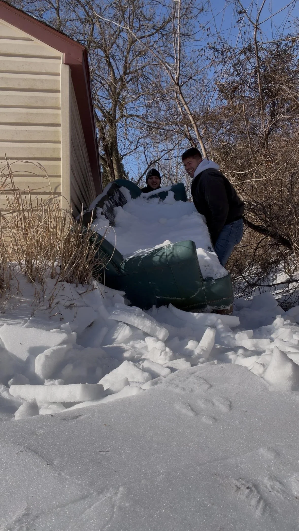 Two young men smiling and playing in the snow with a car that has slid off a house's driveway, surrounded by snow and leafless trees on a clear winter day.