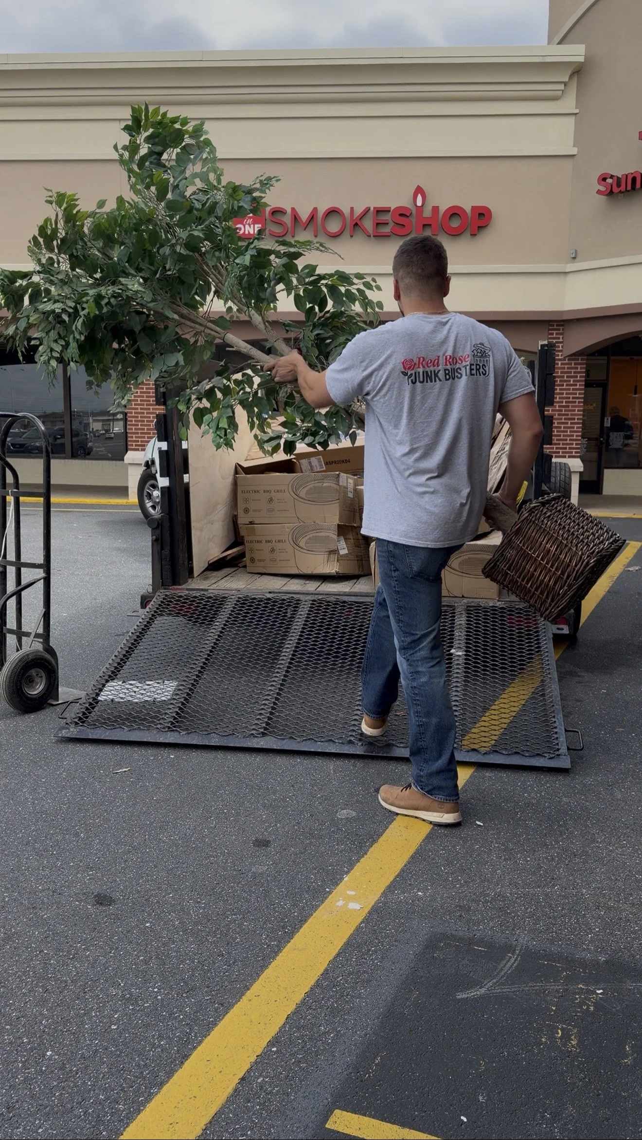 A man unloading a truck outside of a store named 'SMOKESHOP'. He is carrying a small tree or plant and holding a wicker basket. The store has a beige and brick exterior, with part of the store sign visible.