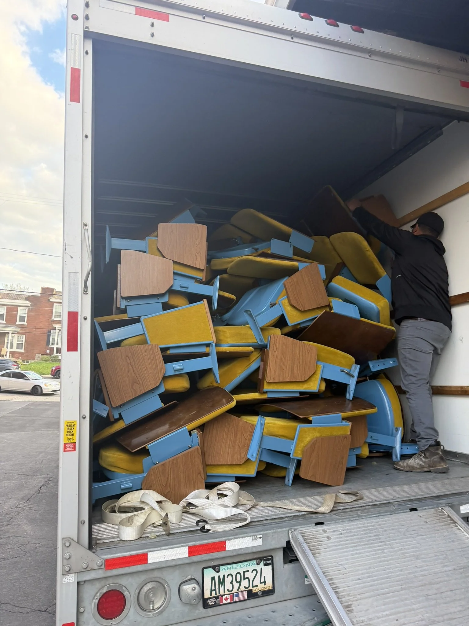 The image shows the back of a moving truck filled with scattered wooden and metal chairs as a person wearing a black jacket and glasses unloads or arranges the chairs inside the truck.
