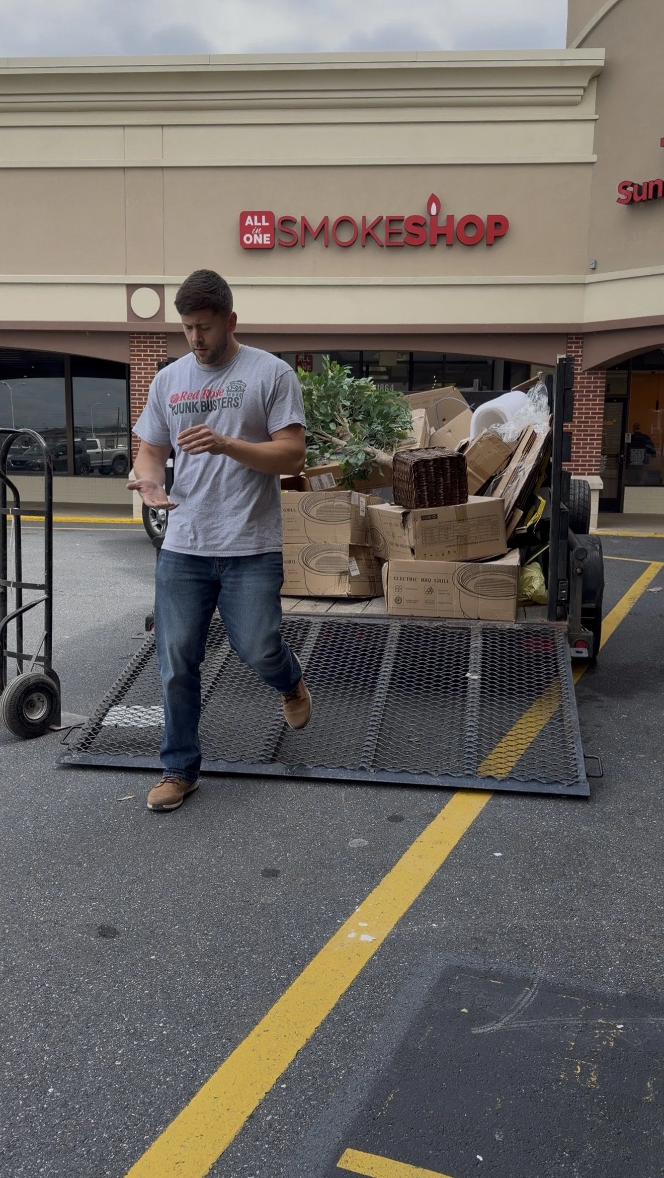 Young man unloading a flatbed trailer full of boxes and goods outside a smoke shop with a parking lot in front.