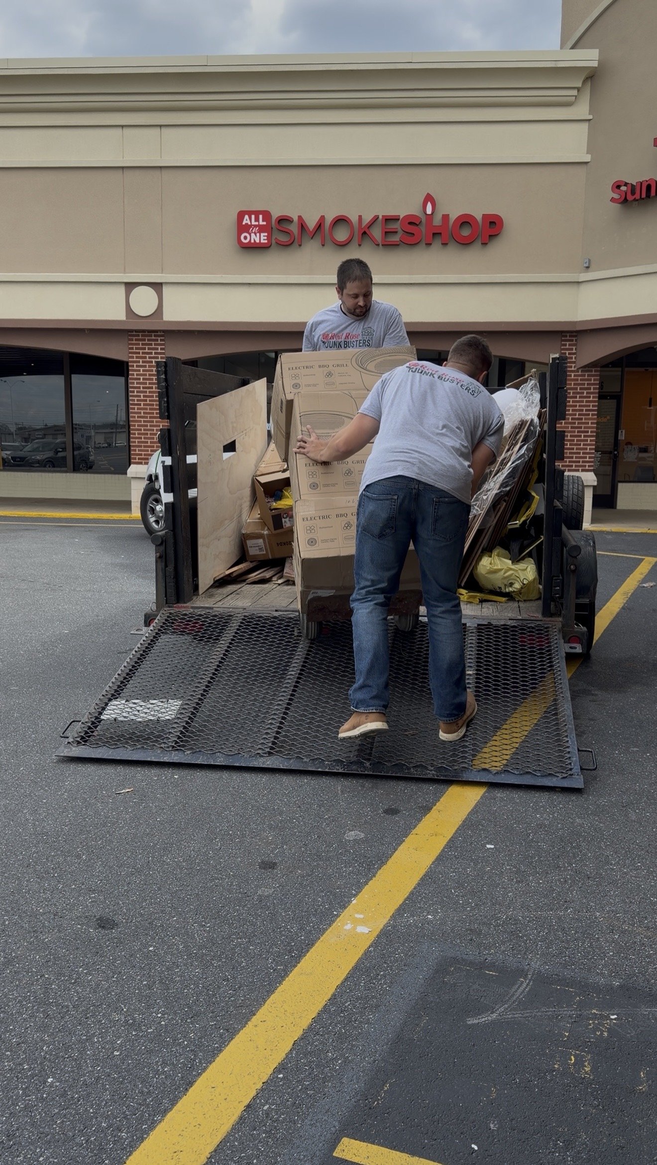Two men loading boxes onto a black flatbed trailer outside a store called Smoke Shop, with a red sign that says "ALL in ONE" and a beige building.