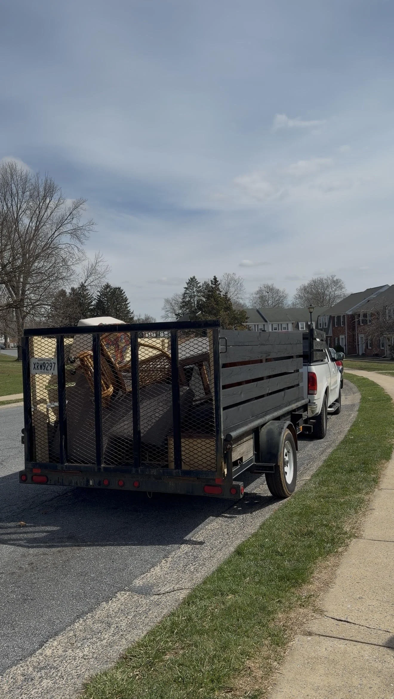 A white pickup truck parked on the side of a residential street with a black utility trailer attached. The trailer is loaded with furniture, including chairs and cardboard boxes. There are houses and trees in the background under a partly cloudy sky.