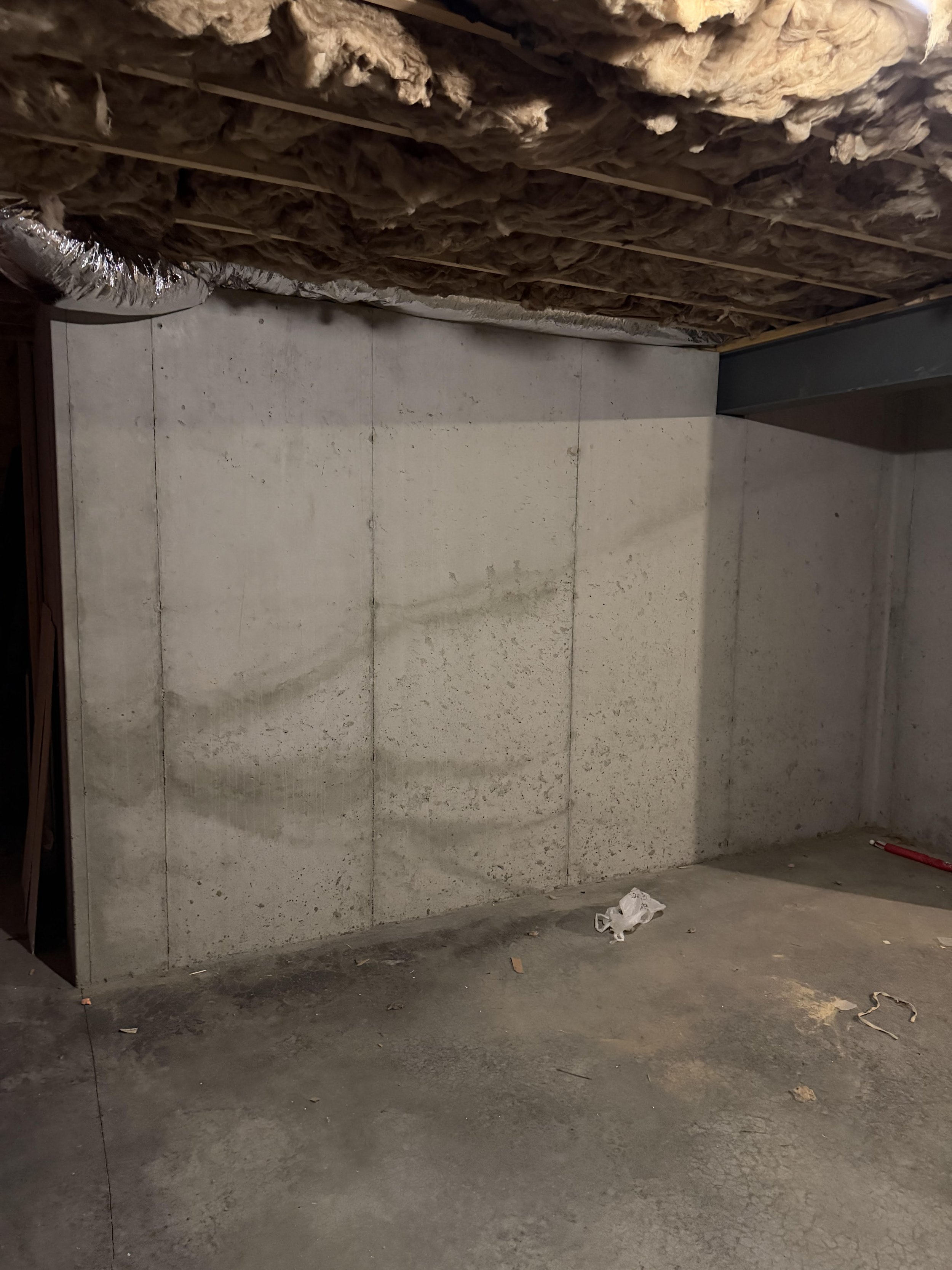 Unfinished basement with concrete floor and concrete wall, insulation exposed on ceiling, and some debris on the floor.