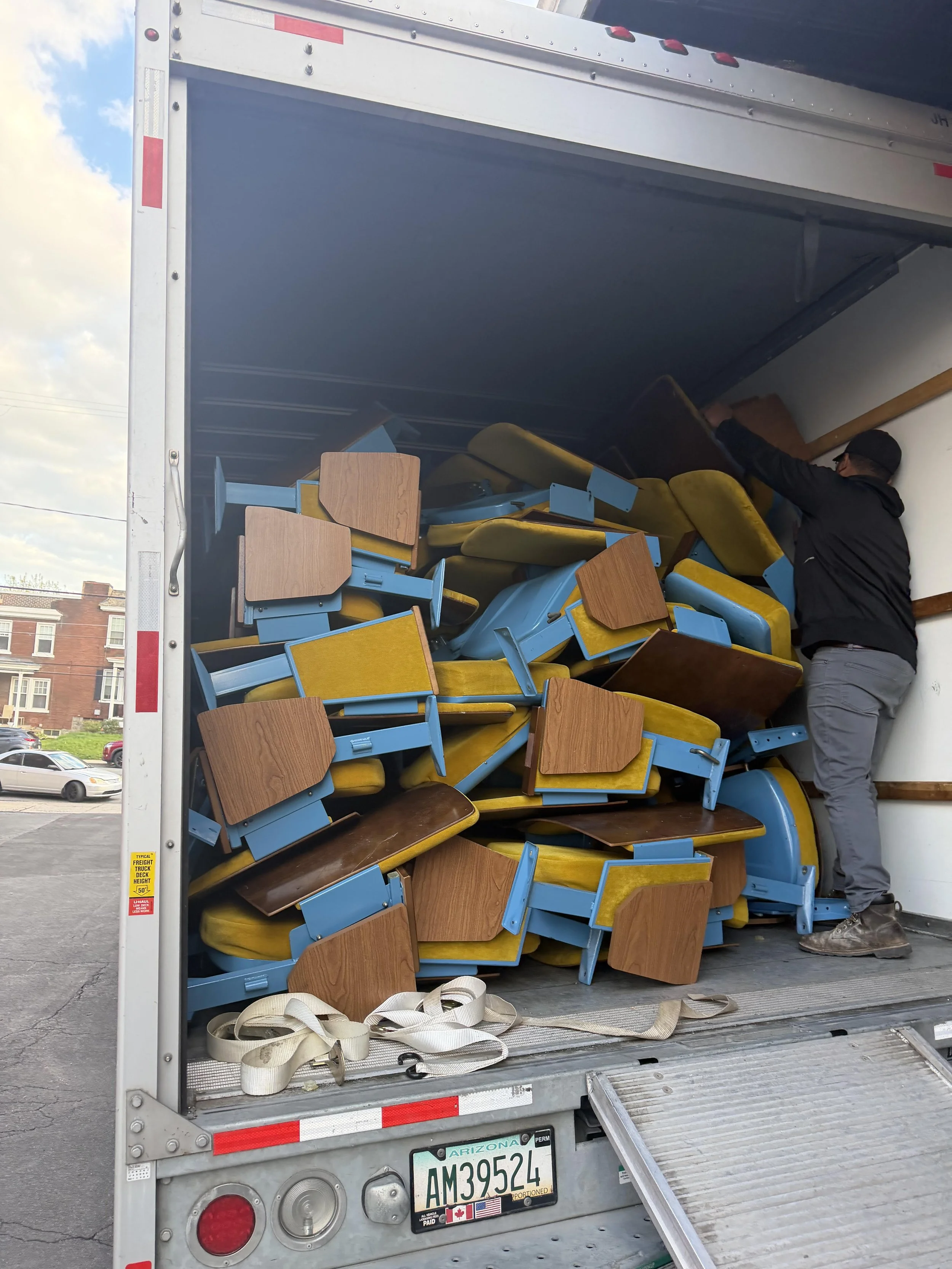 An open delivery truck filled with stacked chairs with wooden seats and blue metal frames, a man in black clothing and glasses loading or unloading the chairs.