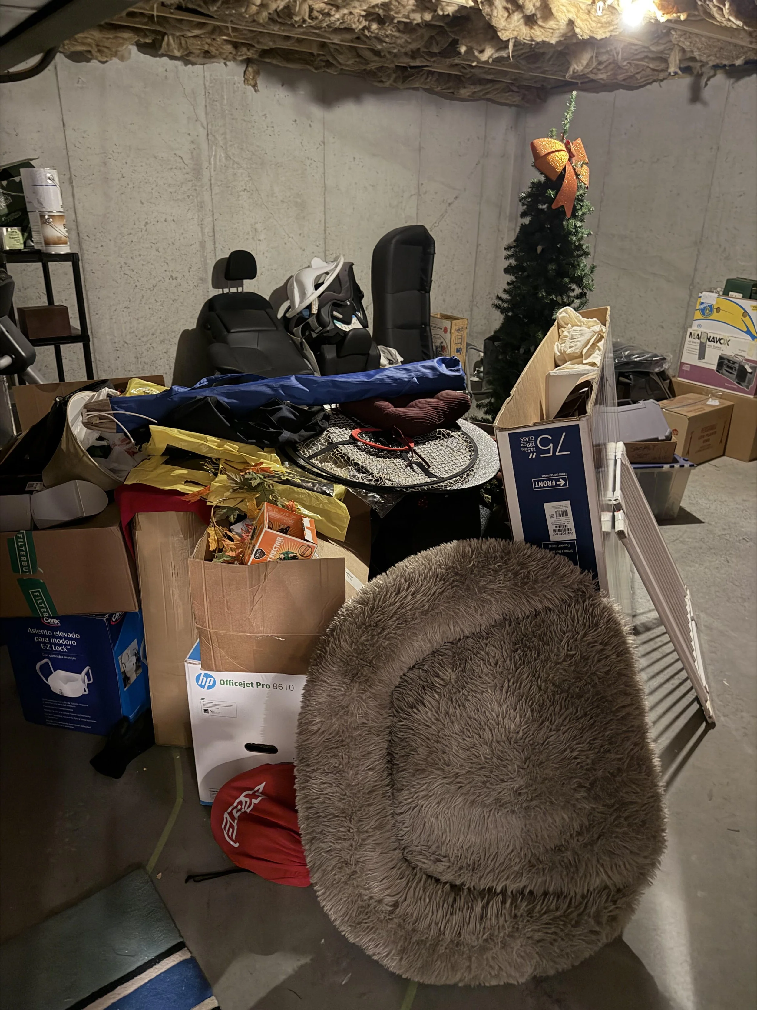A cluttered storage room with various boxes, items, and a small decorated Christmas tree with a large orange bow. There is a fuzzy circular chair in the foreground and a black car seat along with other miscellaneous objects on the table.