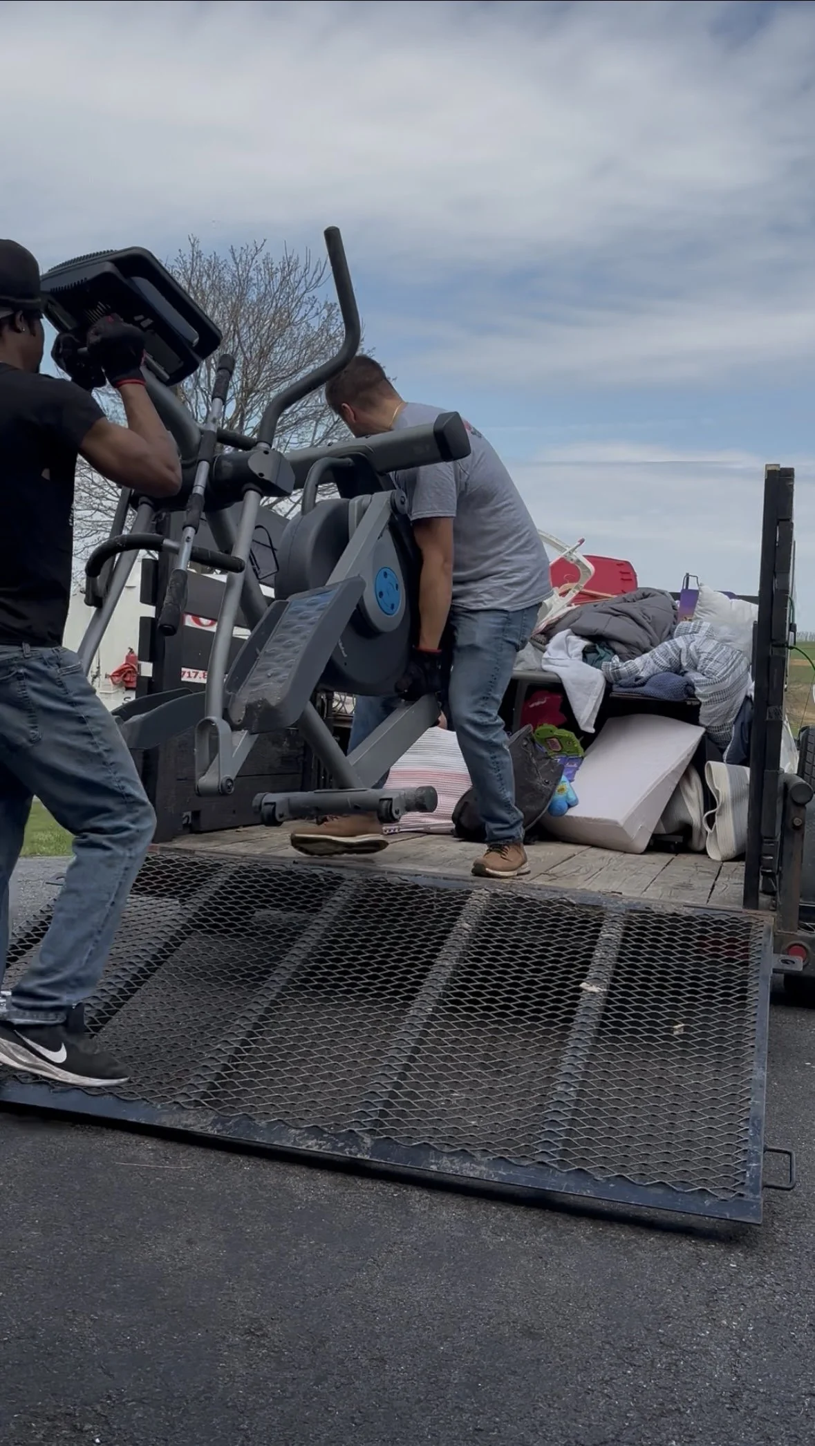 Two men are loading a treadmill onto a flatbed truck with various household items and furniture in the background.