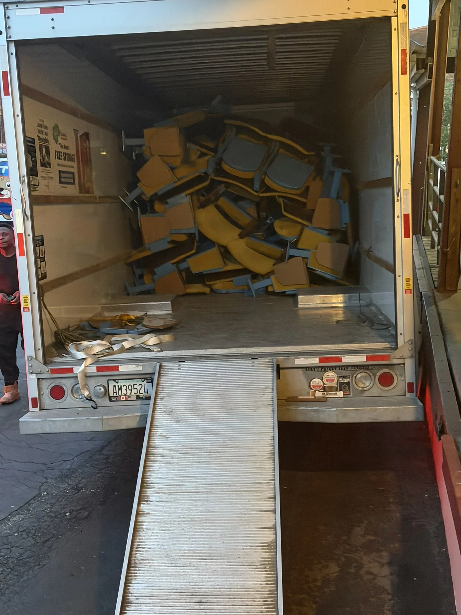 Open truck with beige chairs stacked inside, a metal ramp extending from the back, and items on the floor.