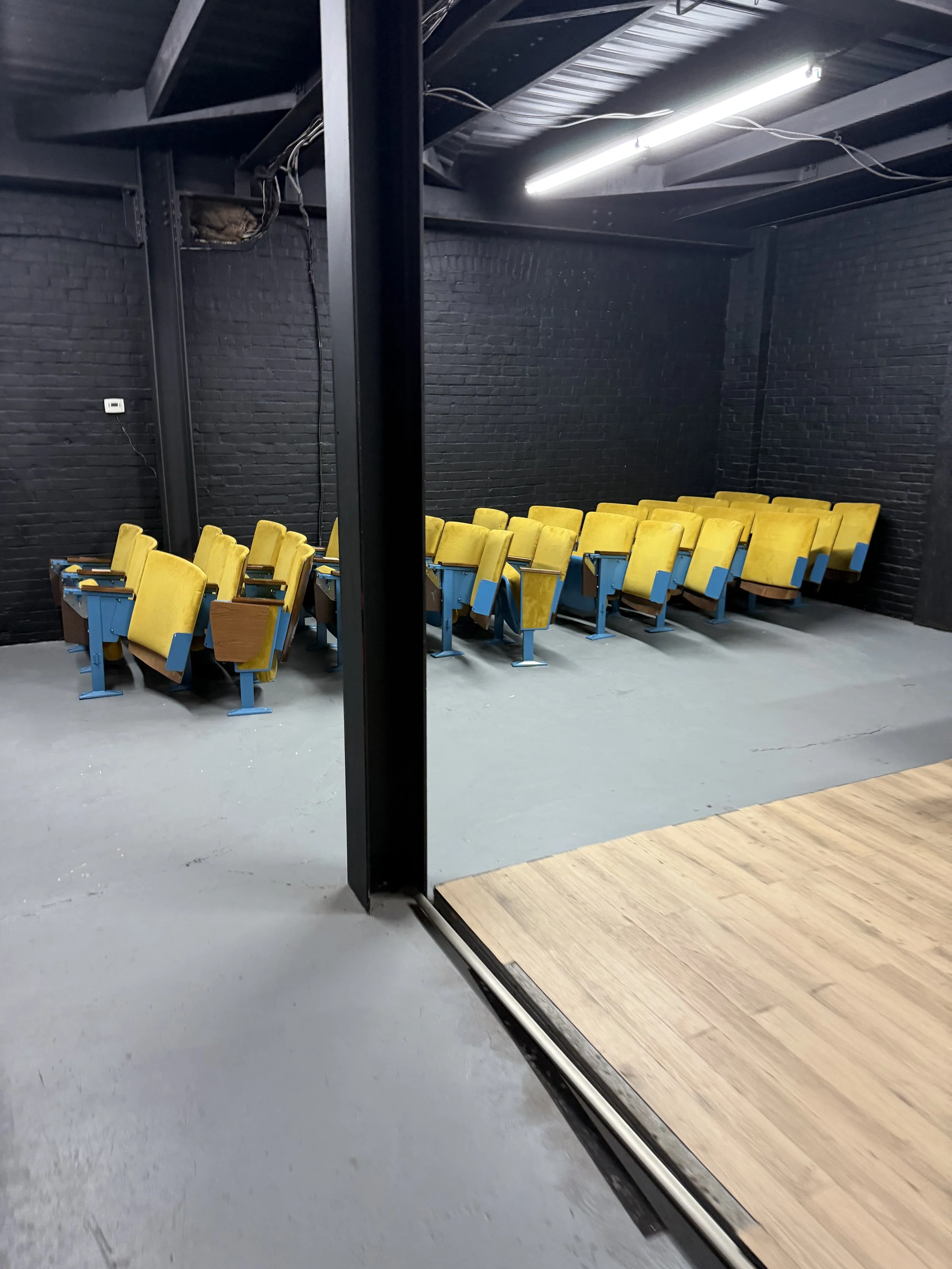 A classroom with yellow chairs arranged in rows, visible through a glass wall with a black frame. The room has black brick walls and a ceiling with exposed beams and a fluorescent light.