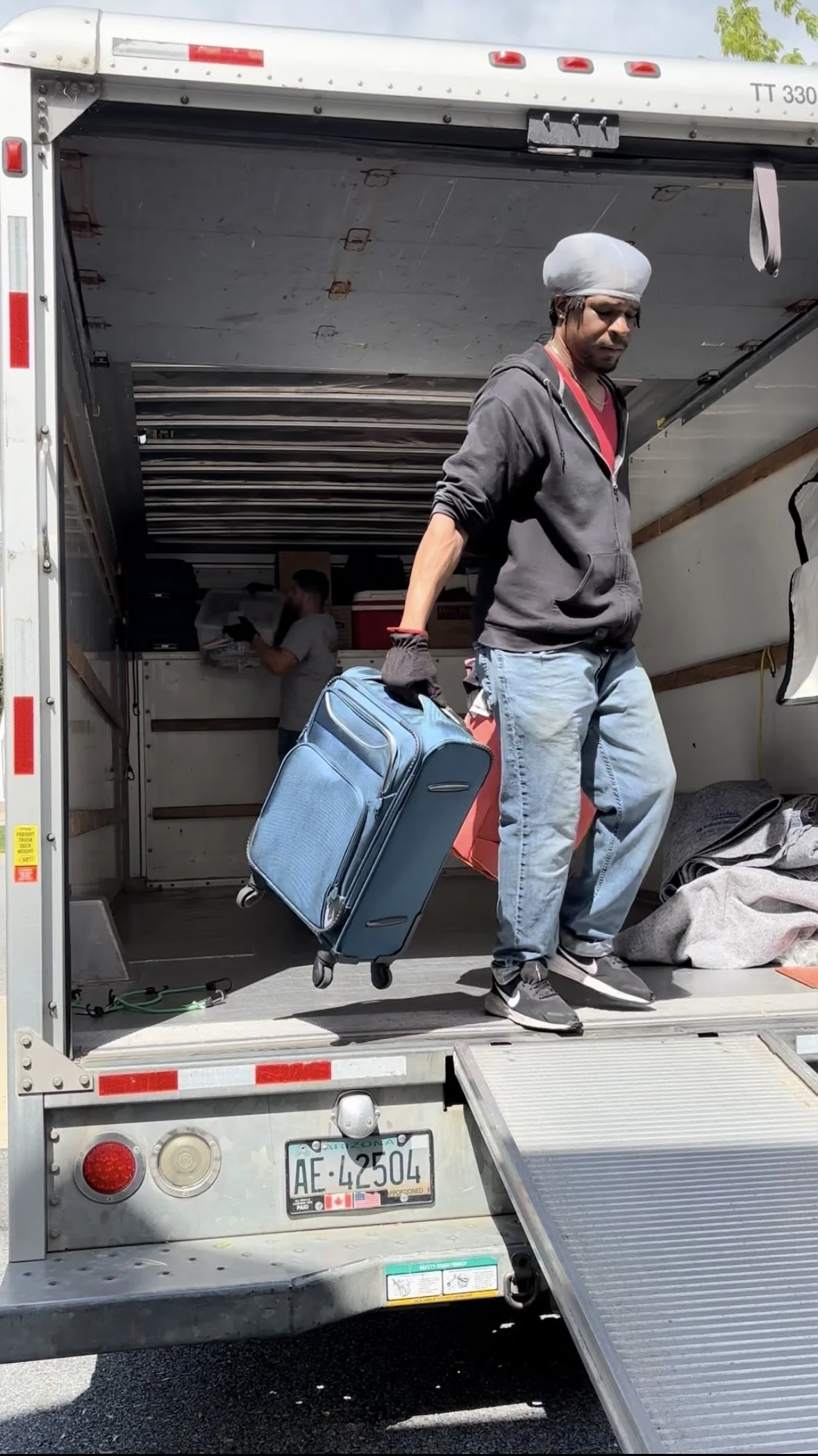 Man loading a blue rolling suitcase into the back of a moving truck, with a woman in the background organizing items inside.