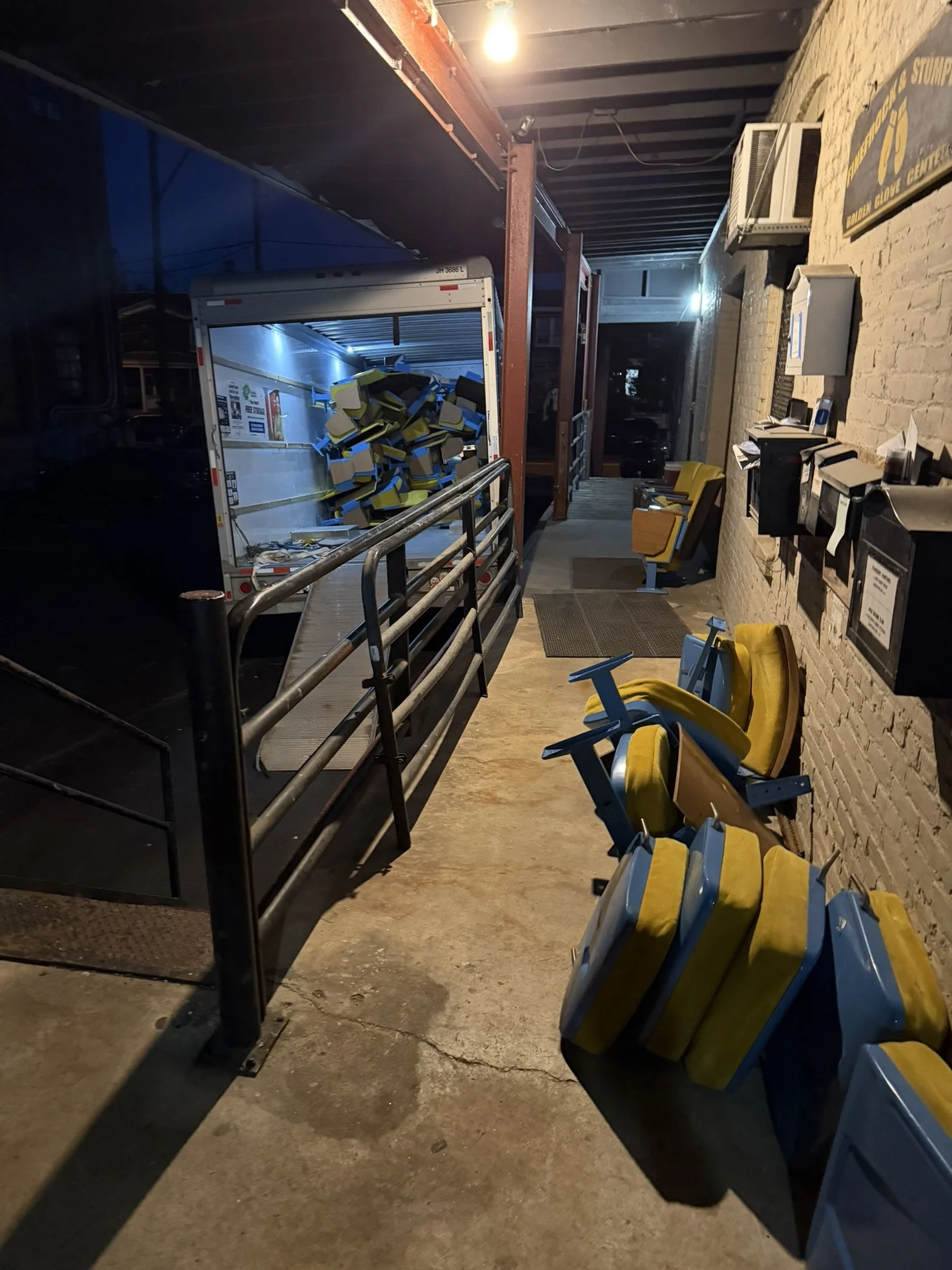 Nighttime scene outside a building with yellow and blue chairs overturned on the ground and a truck with opened doors revealing yellow and black chairs inside. The area is dimly lit with some lights on the wall.