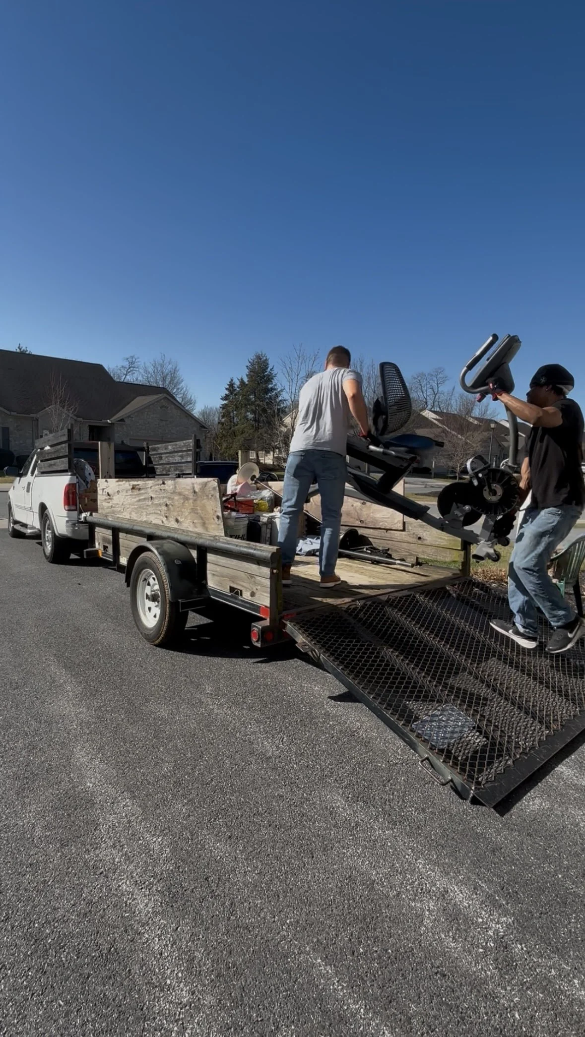 Two men unloading furniture, including an office chair and exercise equipment, from a trailer attached to a pickup truck onto a residential street on a clear, sunny day.