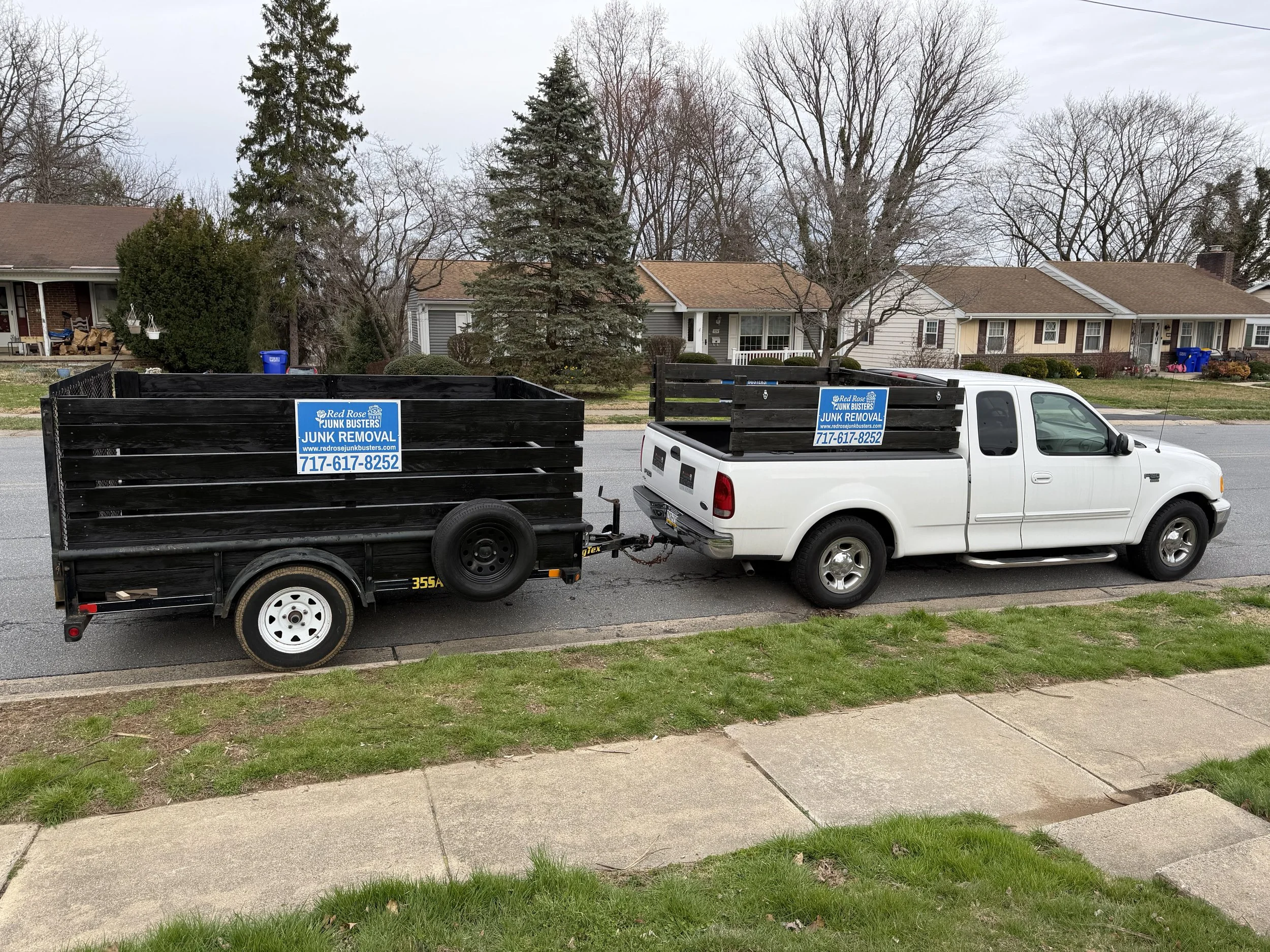 White pickup truck attached to a black trailer with a sign that says 'Junk Removal' on a residential street with houses and trees in the background.
