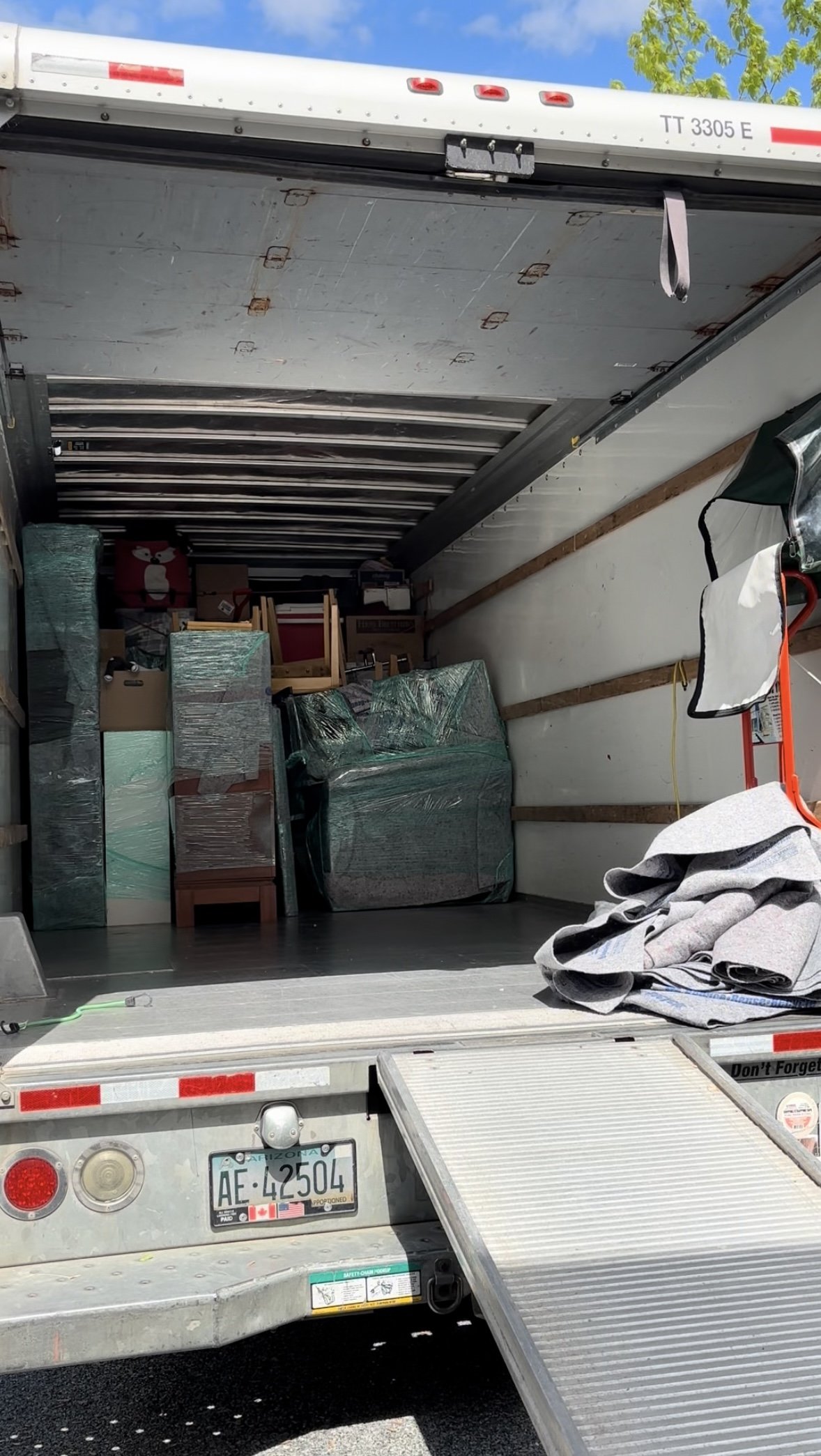 Open moving truck with furniture and boxes inside, some wrapped in plastic, and a skateboard and blanket on the ground at the rear of the truck.