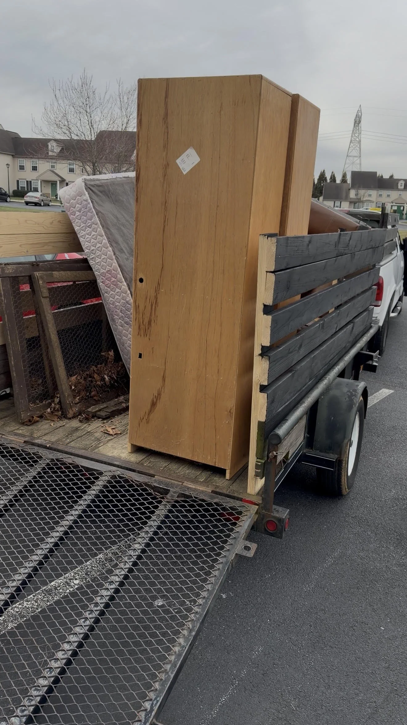 A trailer loaded with furniture and a mattress parked in a parking lot, with residential houses and trees in the background.