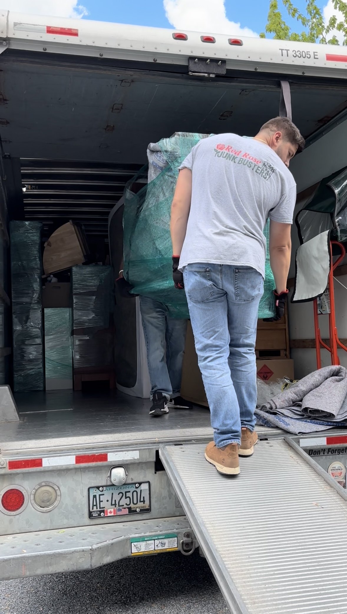 People loading furniture and boxes into a moving truck with a ramp, parked on a sunny day.