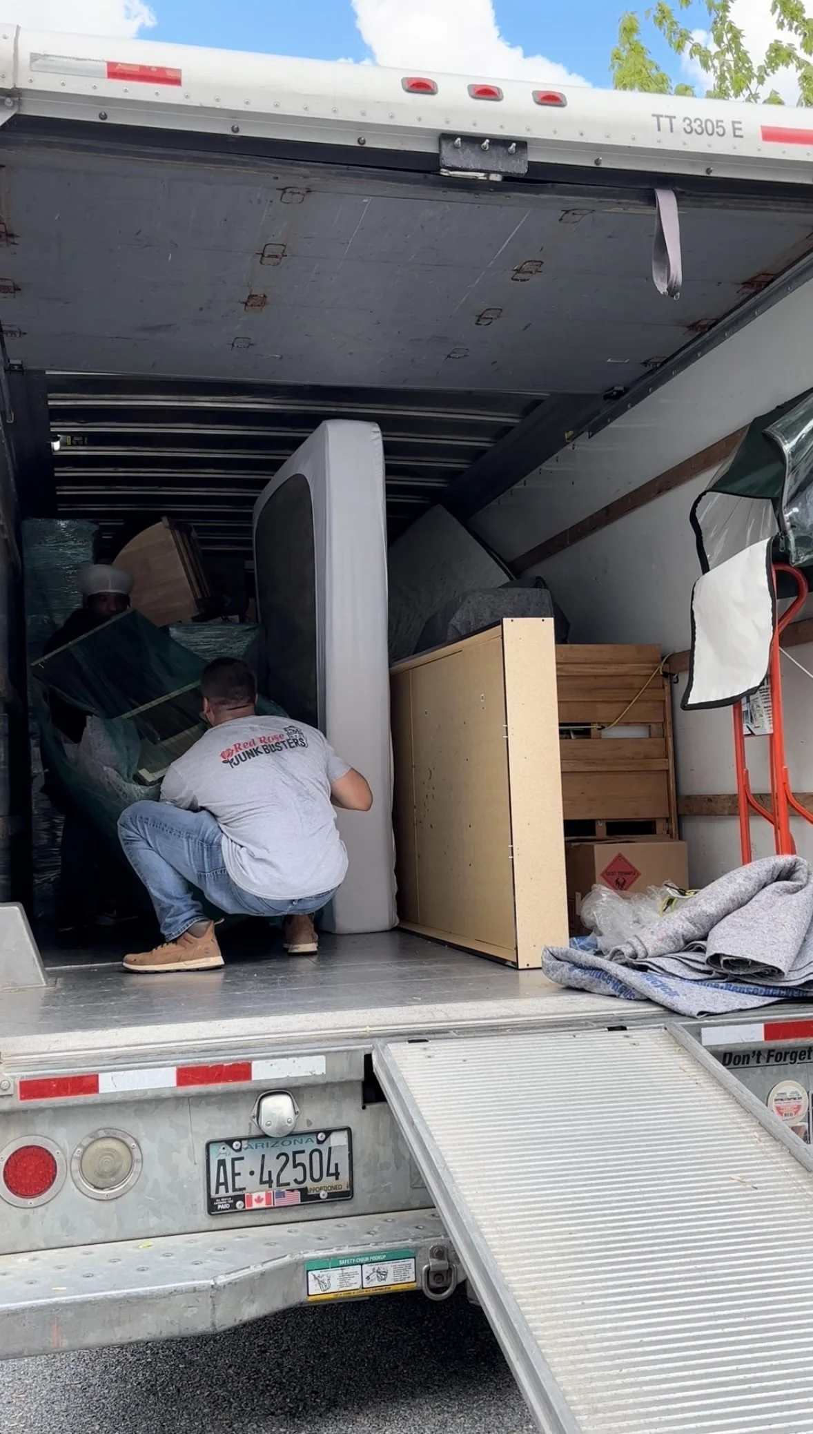 People loading furniture and boxes into a moving truck with an Arizona license plate, under a blue sky with some clouds.