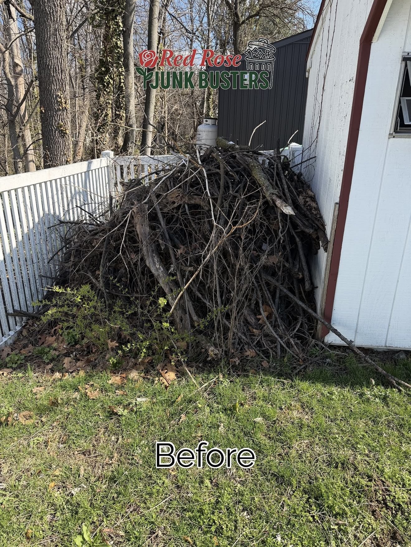 Pile of dead branches and wood next to a white fence and a building with a sign that reads 'Red Rose Junk Busters'.