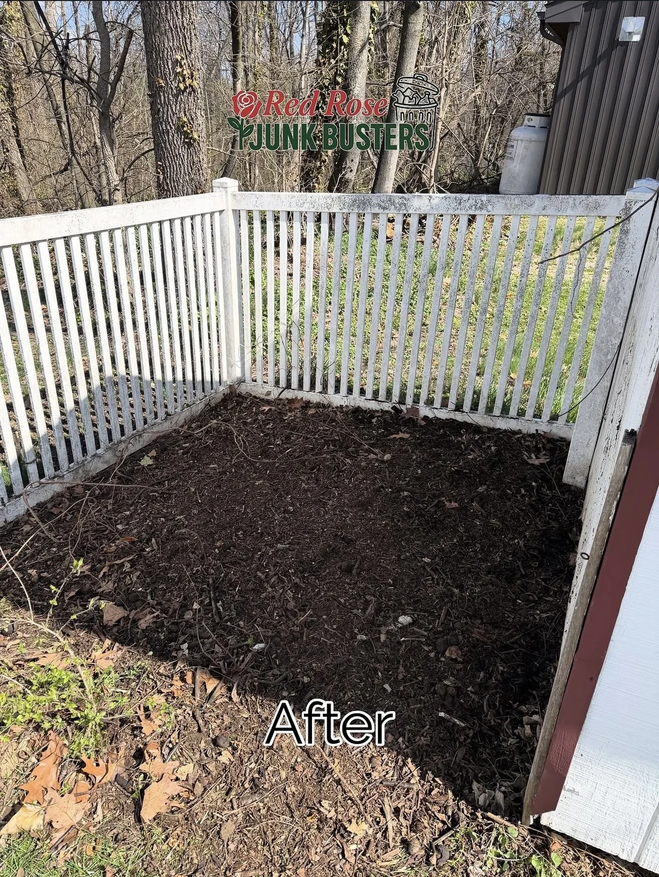 Cleaned and prepared backyard garden bed enclosed by white rail fence, with a mulch layer on the ground and some leaves. Trees and a small building are visible in the background. Sign for 'Red Rose Junk Busters' is in the background.
