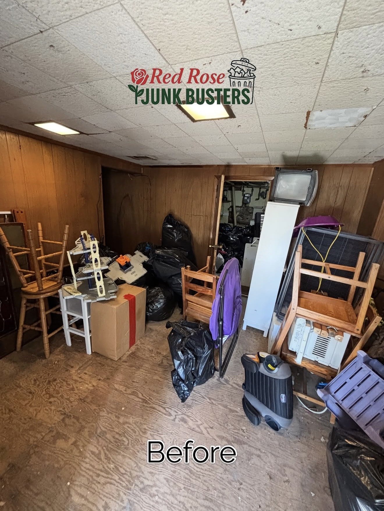 Room filled with junk including stacked chairs, black trash bags, a cardboard box, a purple backpack, and miscellaneous household items, with a wooden wall and tiled ceiling.