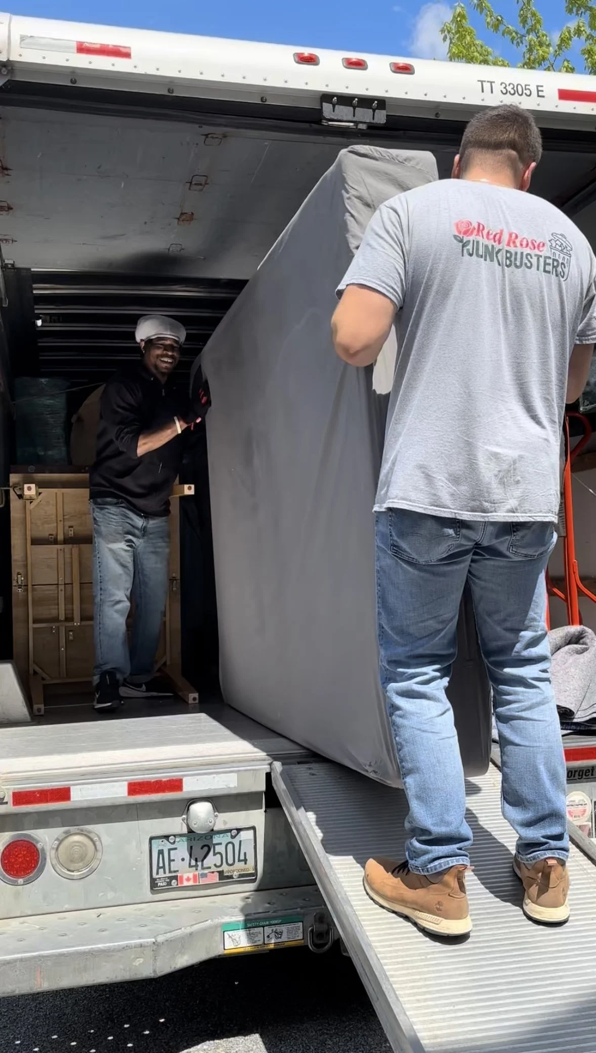 Two men loading furniture onto a moving truck. One man is wearing a gray 'Red Rose Junk Busters' T-shirt and tan shoes. The other man, wearing a hat and gloves, is smiling as he helps. The truck has a ramp and a box truck registration plate from Ariz