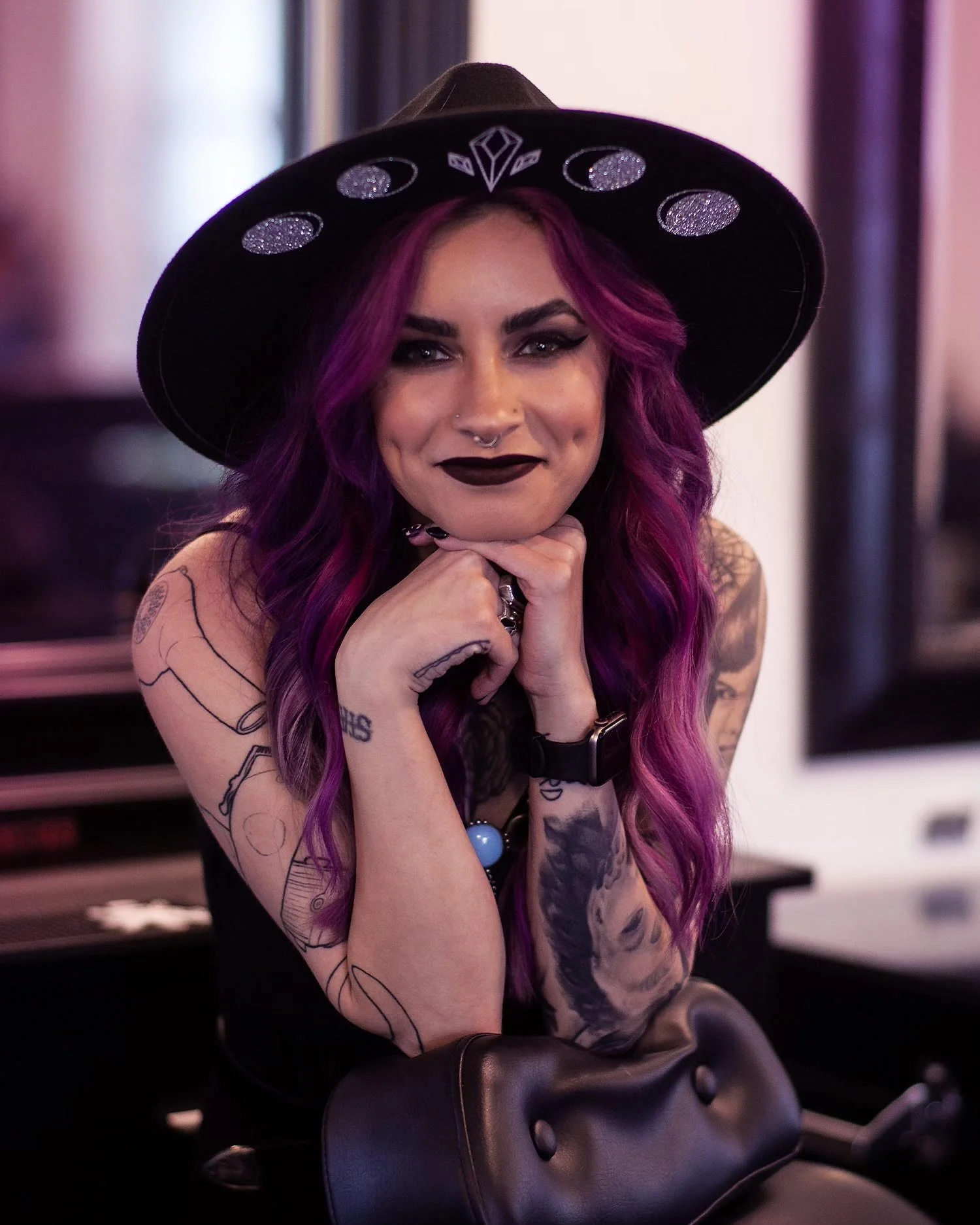 A woman with long purple hair wearing a black hat with glittery designs, black lipstick, tattoos on her arms, and a nose piercing, resting her chin on her hands and smiling.
