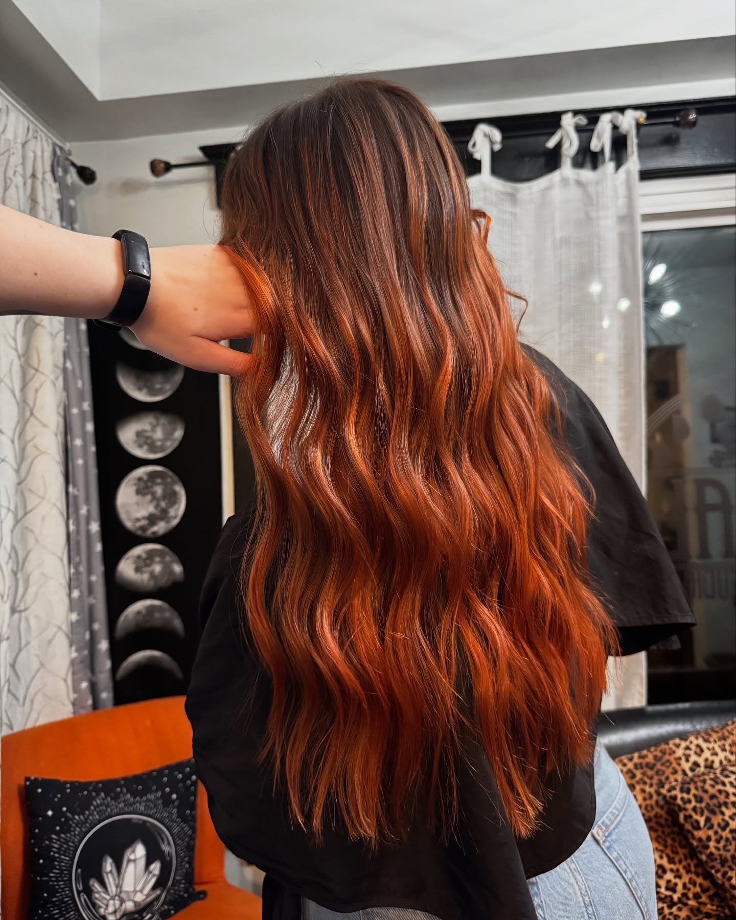 Back view of a woman with long, wavy, reddish-orange hair in a cozy room with moon phase decor on the wall and animal print decor on a chair.