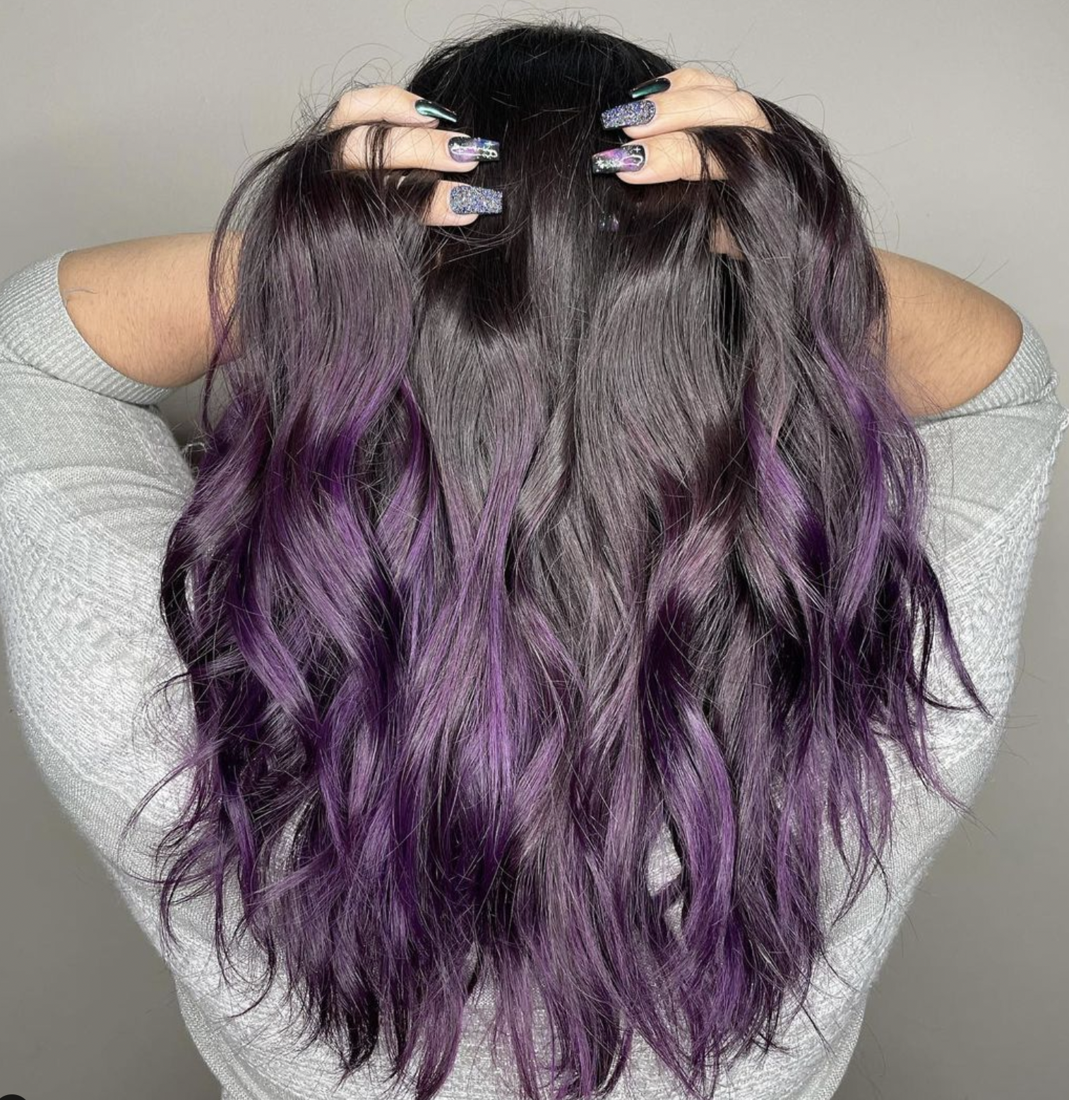 Person with long, wavy hair that transitions from dark gray at the roots to purple at the tips, holding their head with both hands, showing manicured nails with glittery, colorful designs.