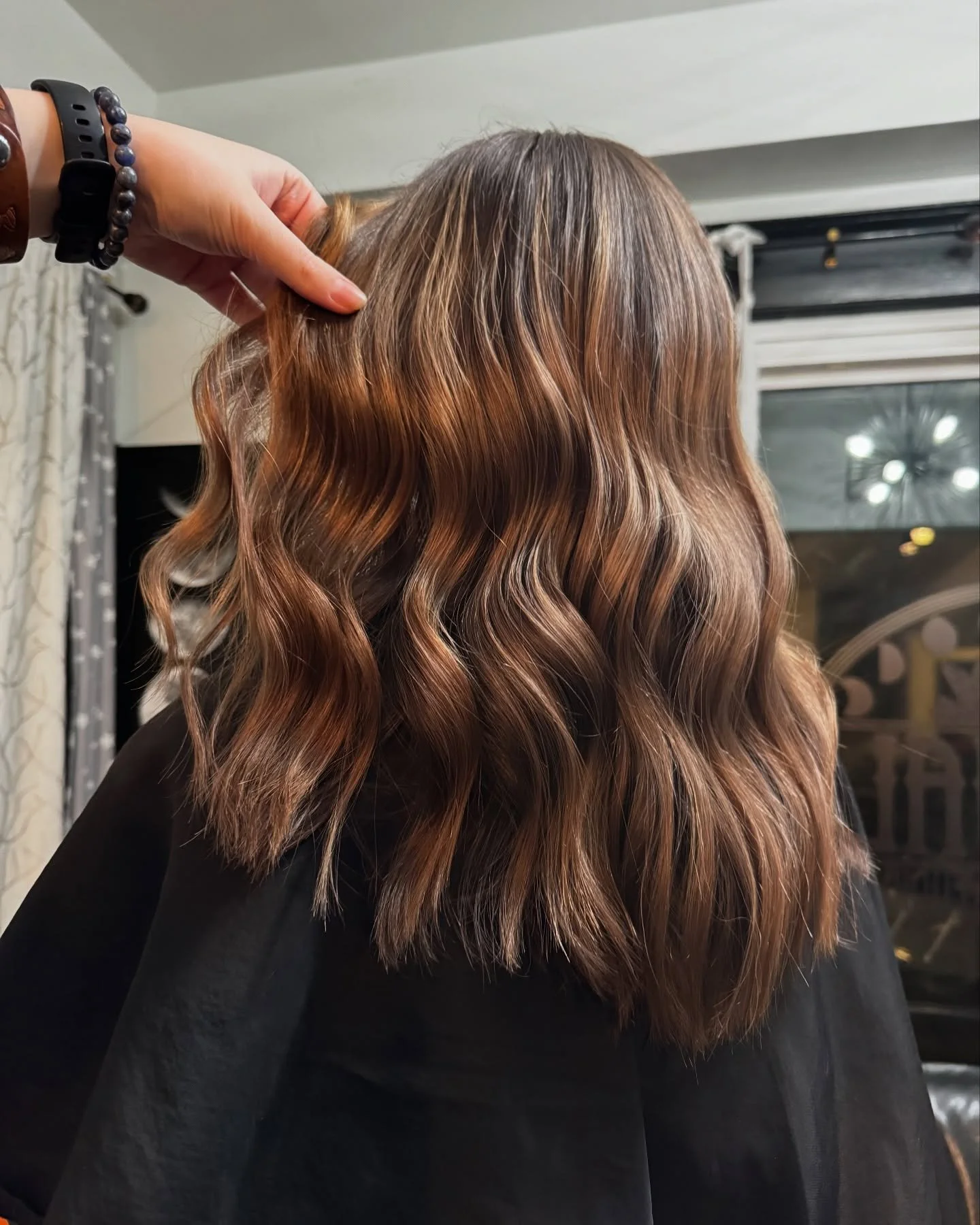 Back of a woman with wavy, shoulder-length brown hair with caramel highlights, sitting in a salon chair.