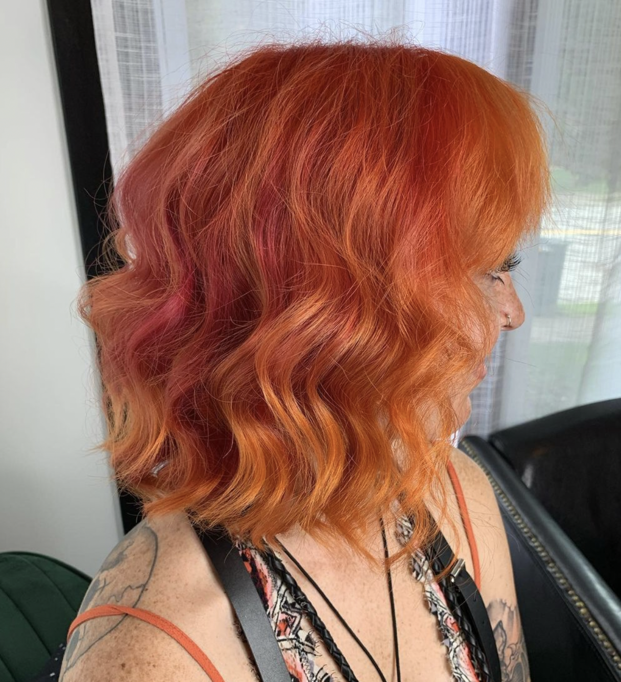Side view of a woman with orange-red, wavy hair, wearing a sleeveless top, tattoos on her shoulder, and a nose ring, sitting indoors near a window with curtains.