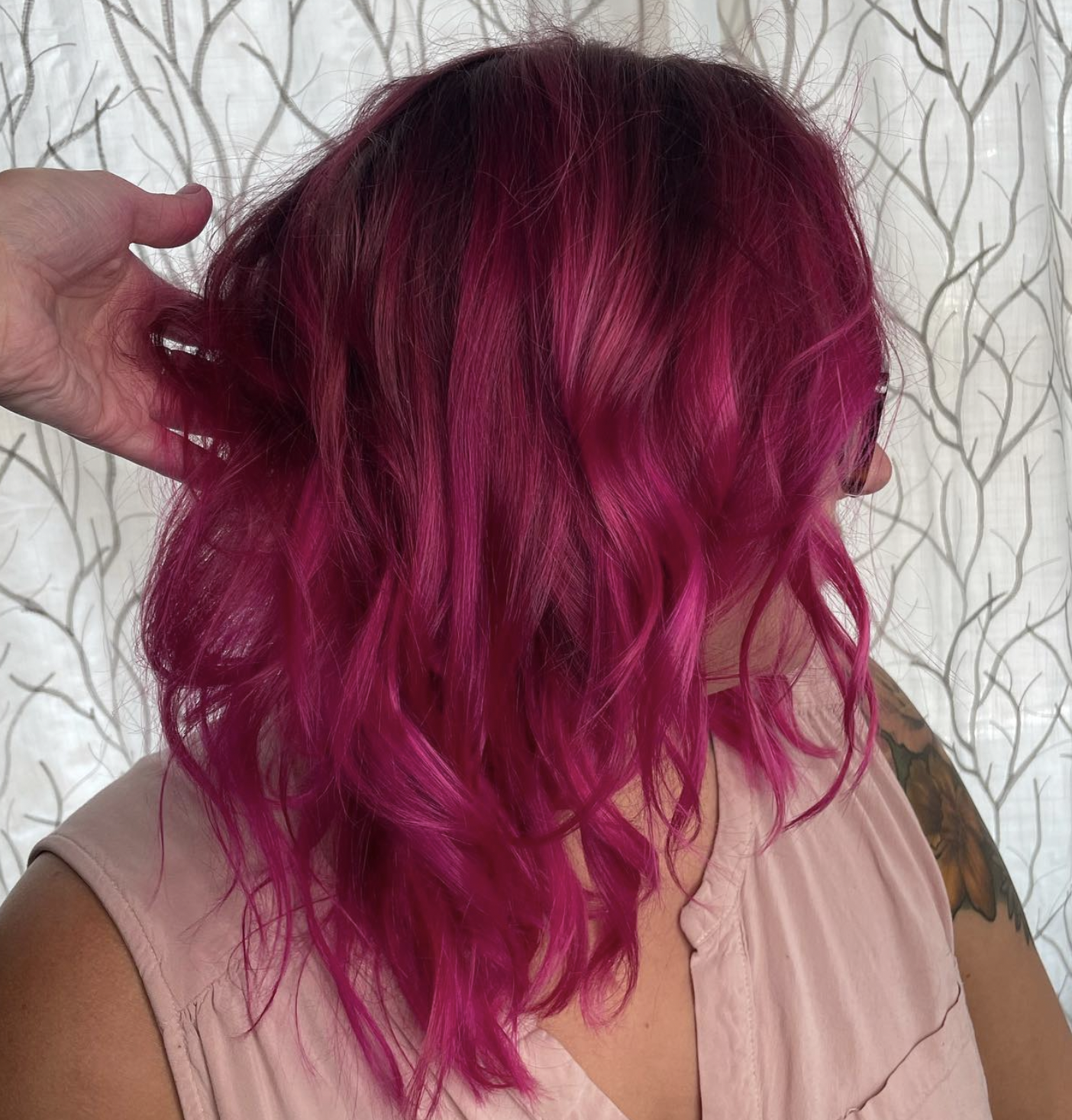 Woman with vibrant hot pink, wavy hair, partially covered face, wearing a sleeveless pink top, standing in front of a light-colored curtain with a leafy tree pattern.