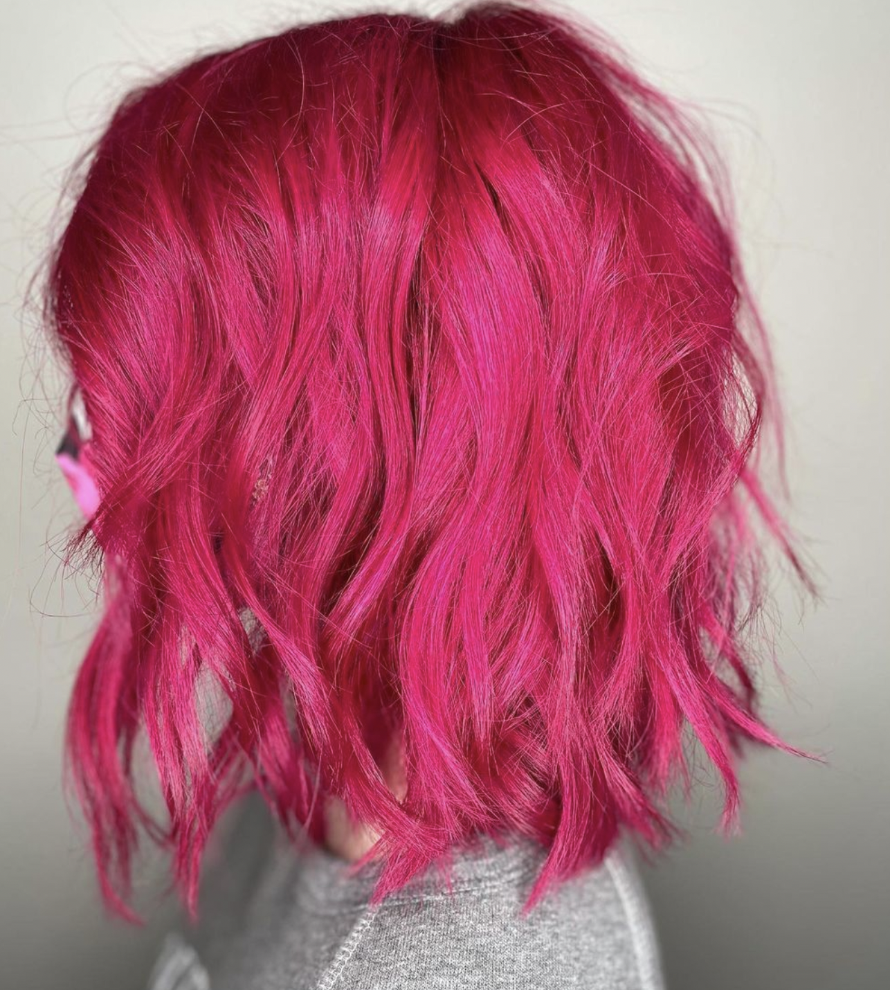 Back view of a person with shoulder-length, wavy, bright pink hair wearing a gray shirt.