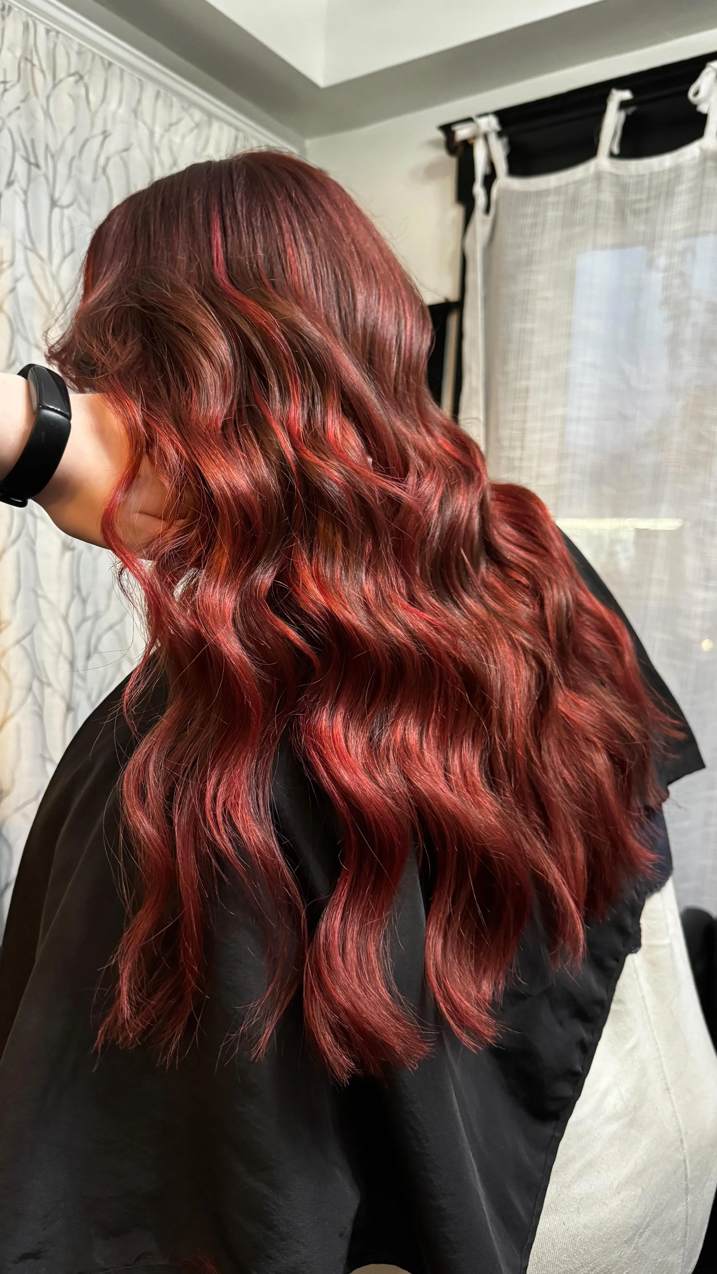 A woman with long, wavy, red hair sitting in a salon chair, wearing a black cape.