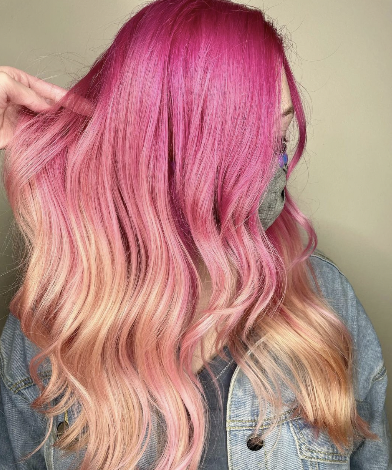 Woman with long, wavy hair dyed in shades of pink and peach, wearing a face mask and denim jacket.