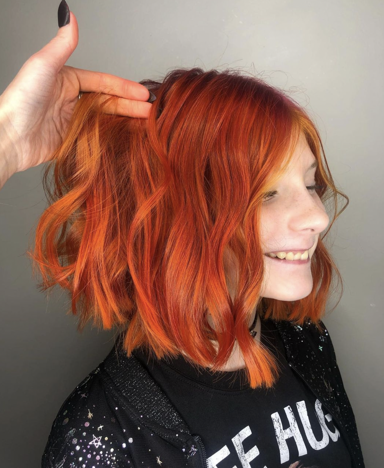 A woman with bright red, wavy hair smiling and looking to her right, with a person's hand holding her hair from behind.