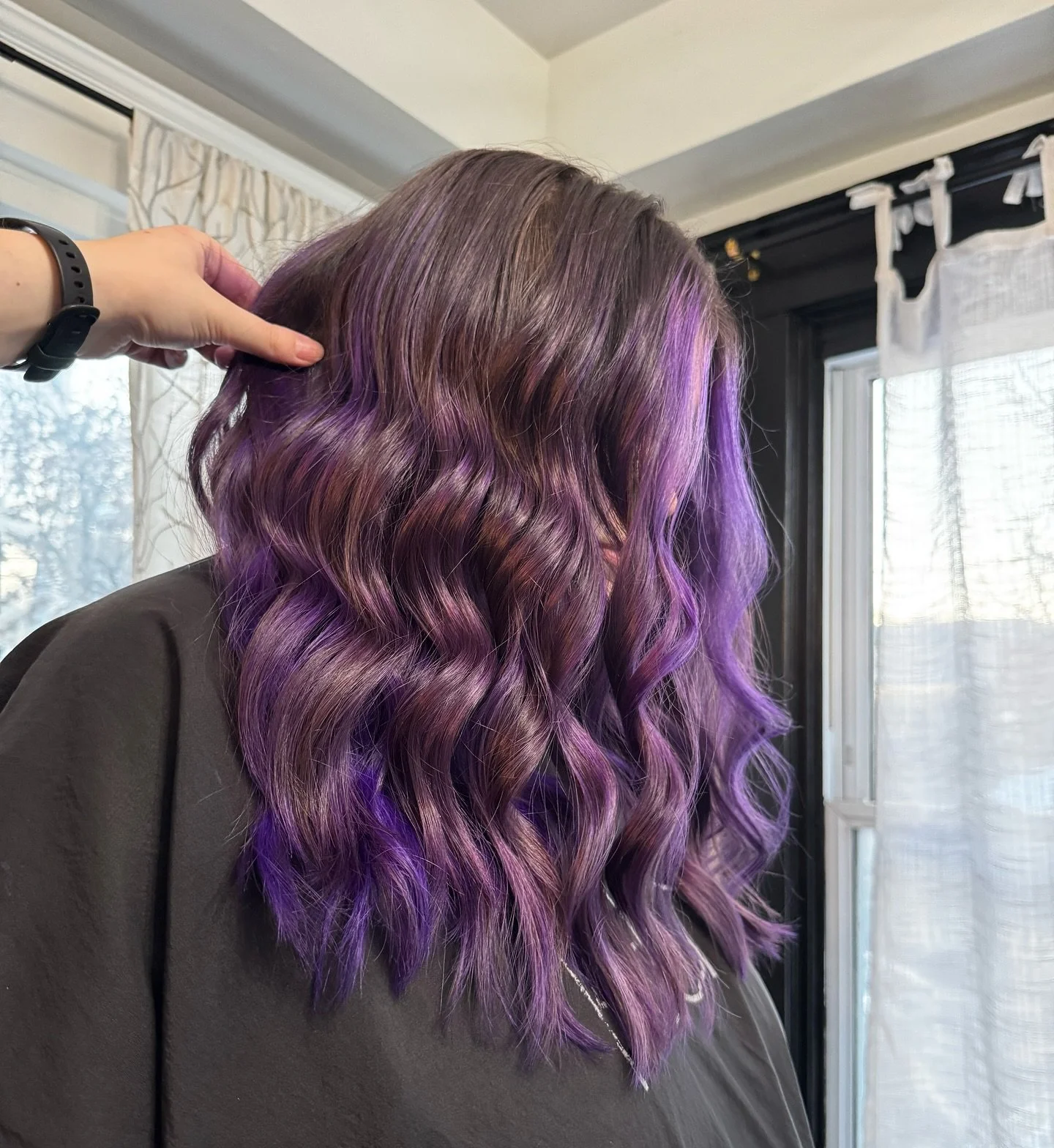 Person with wavy hair dyed in shades of purple and lavender, being styled in a room with curtains and a window in the background.