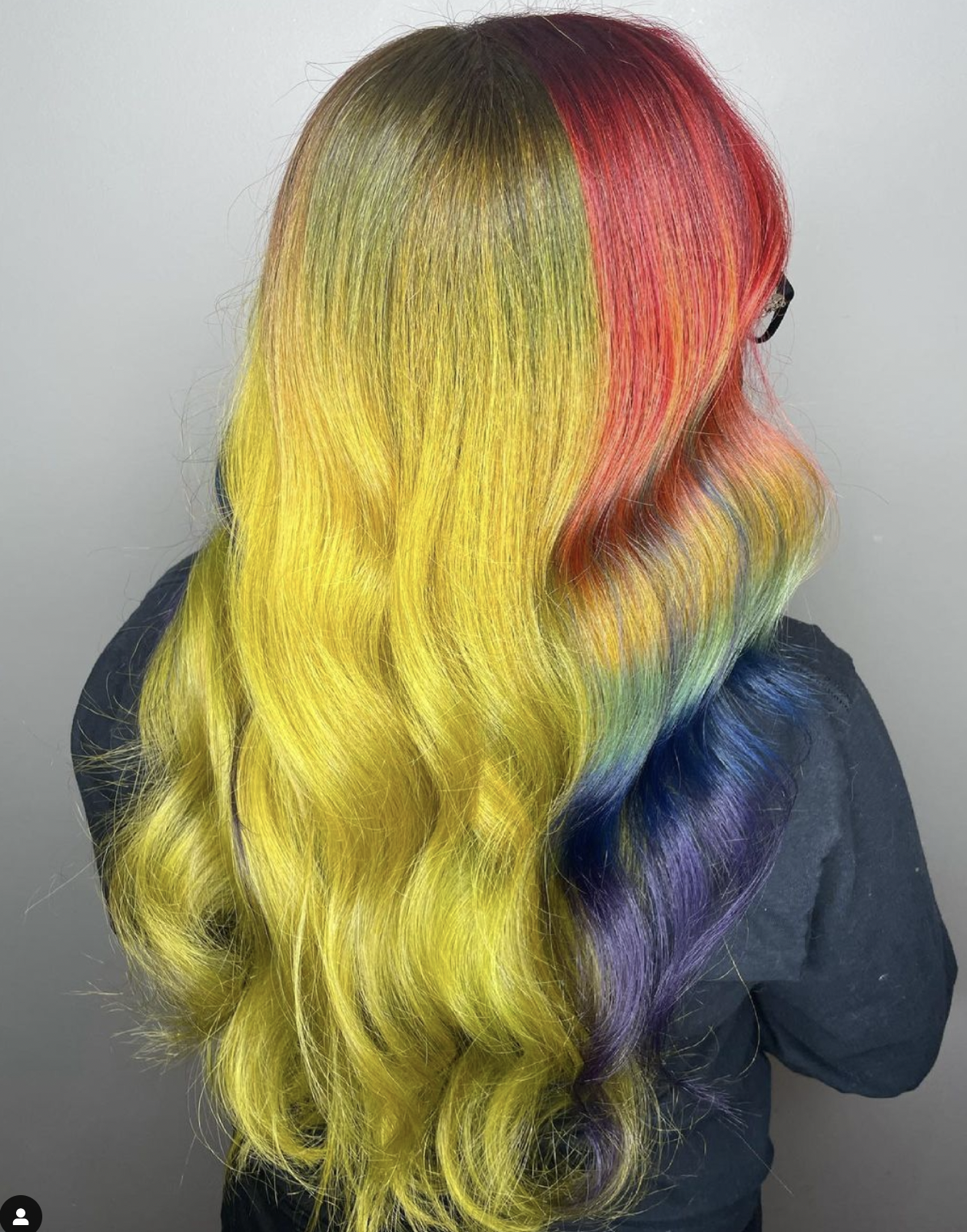 Back view of a person with long, wavy rainbow-colored hair, including red, orange, yellow, green, blue, and purple.