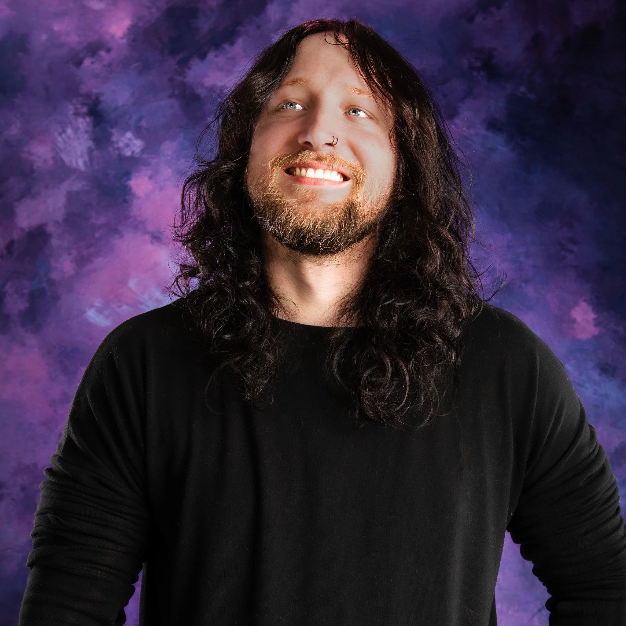 A smiling man with long, curly dark hair, a nose piercing, a beard, wearing a black shirt, against a purple and blue mottled background.