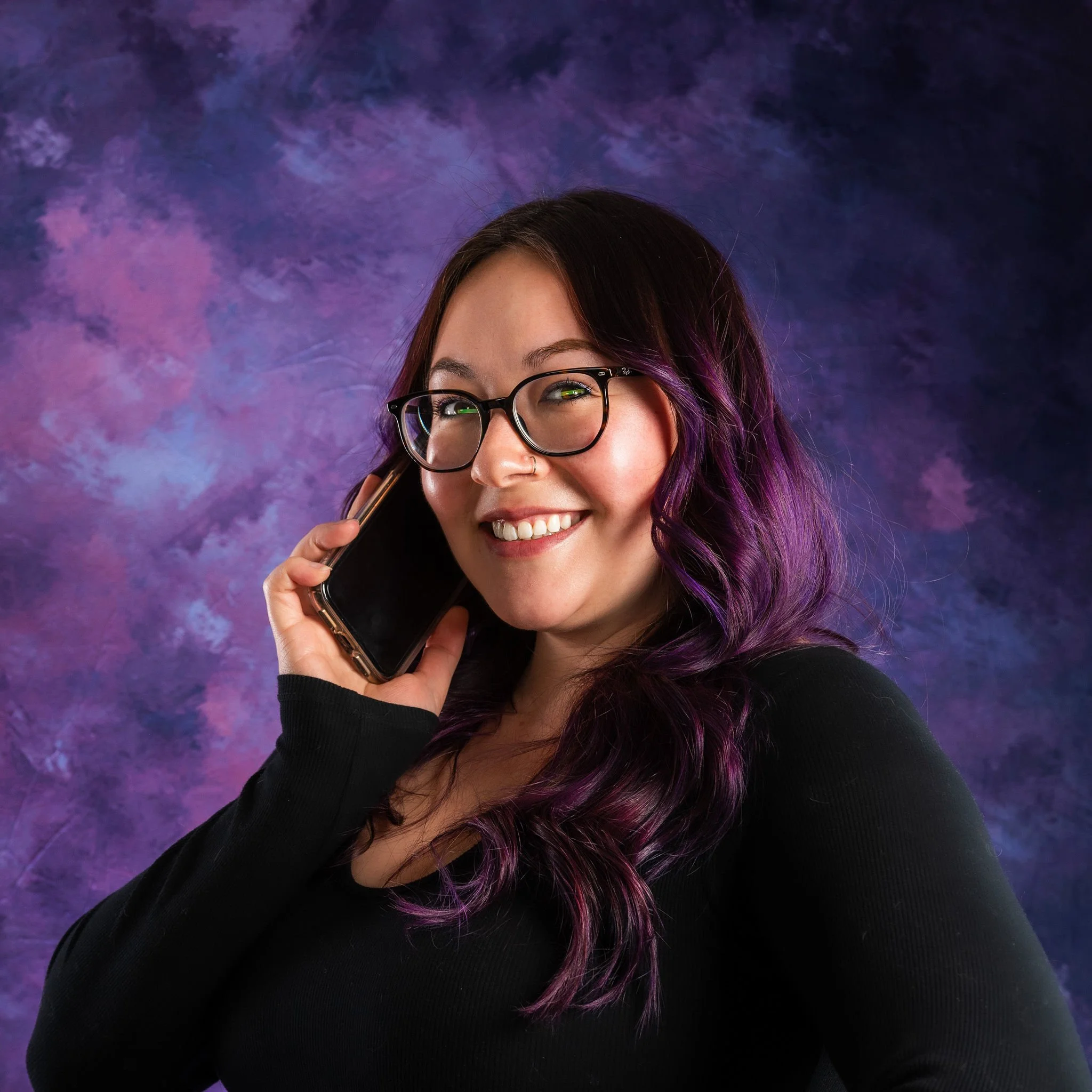A woman with dark purple hair and glasses smiling while talking on her cellphone against a purple and blue textured background.