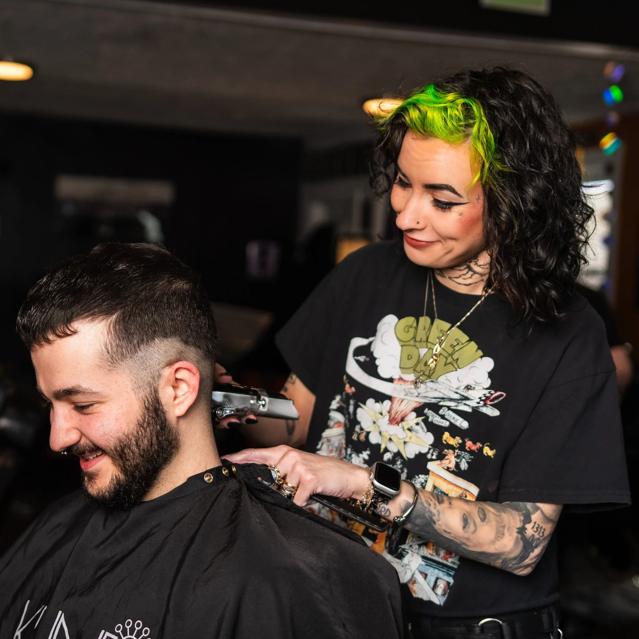 A woman with dyed green hair and tattoos is giving a man a haircut in a barbershop. The woman is smiling and looking down at the man, who is sitting in a barber chair wearing a black cape.