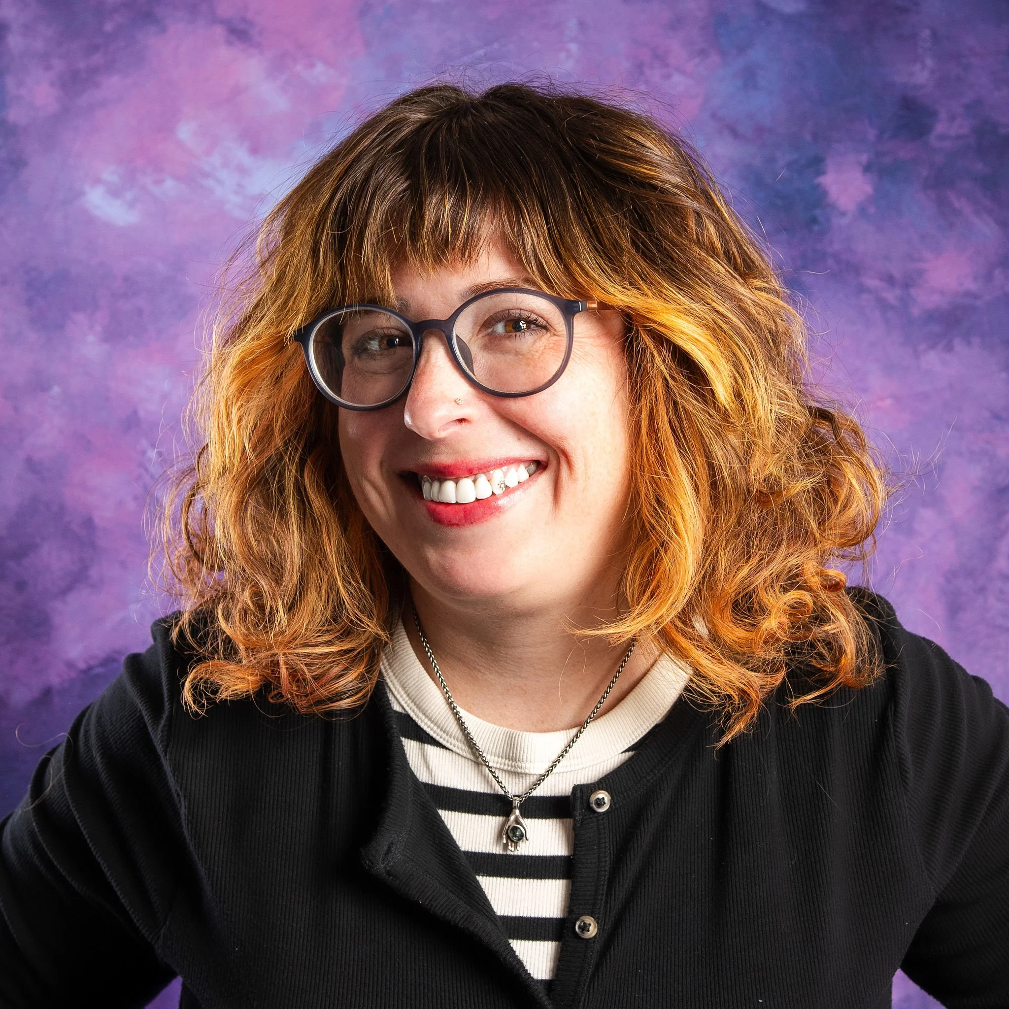 A woman with curly red hair, wearing glasses, a striped shirt, and a necklace, smiling against a purple and blue watercolor background.