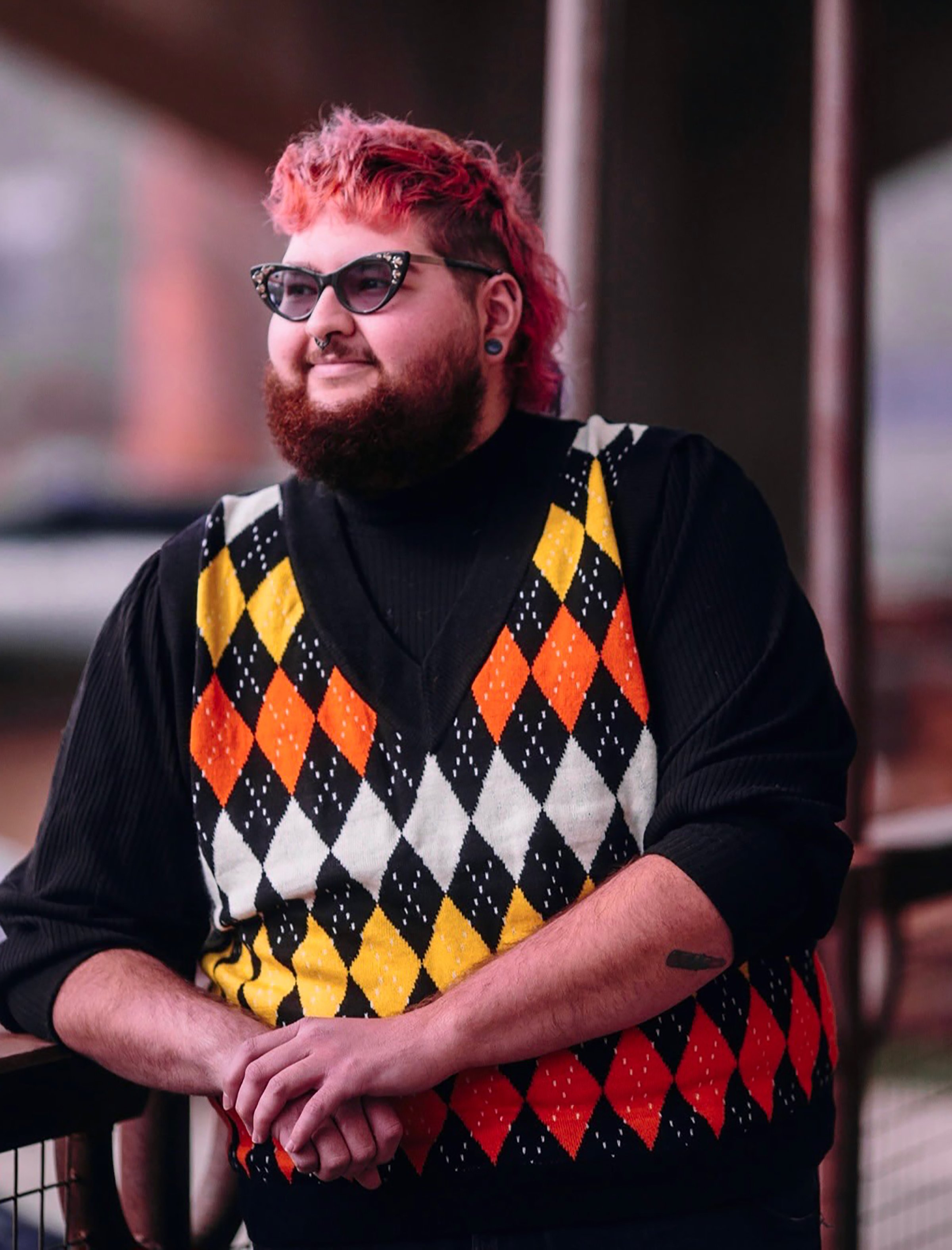 A bearded man with pink hair and black Kaiser sunglasses, wearing a black sweater and a colorful argyle vest, leaning on a railing outdoors.