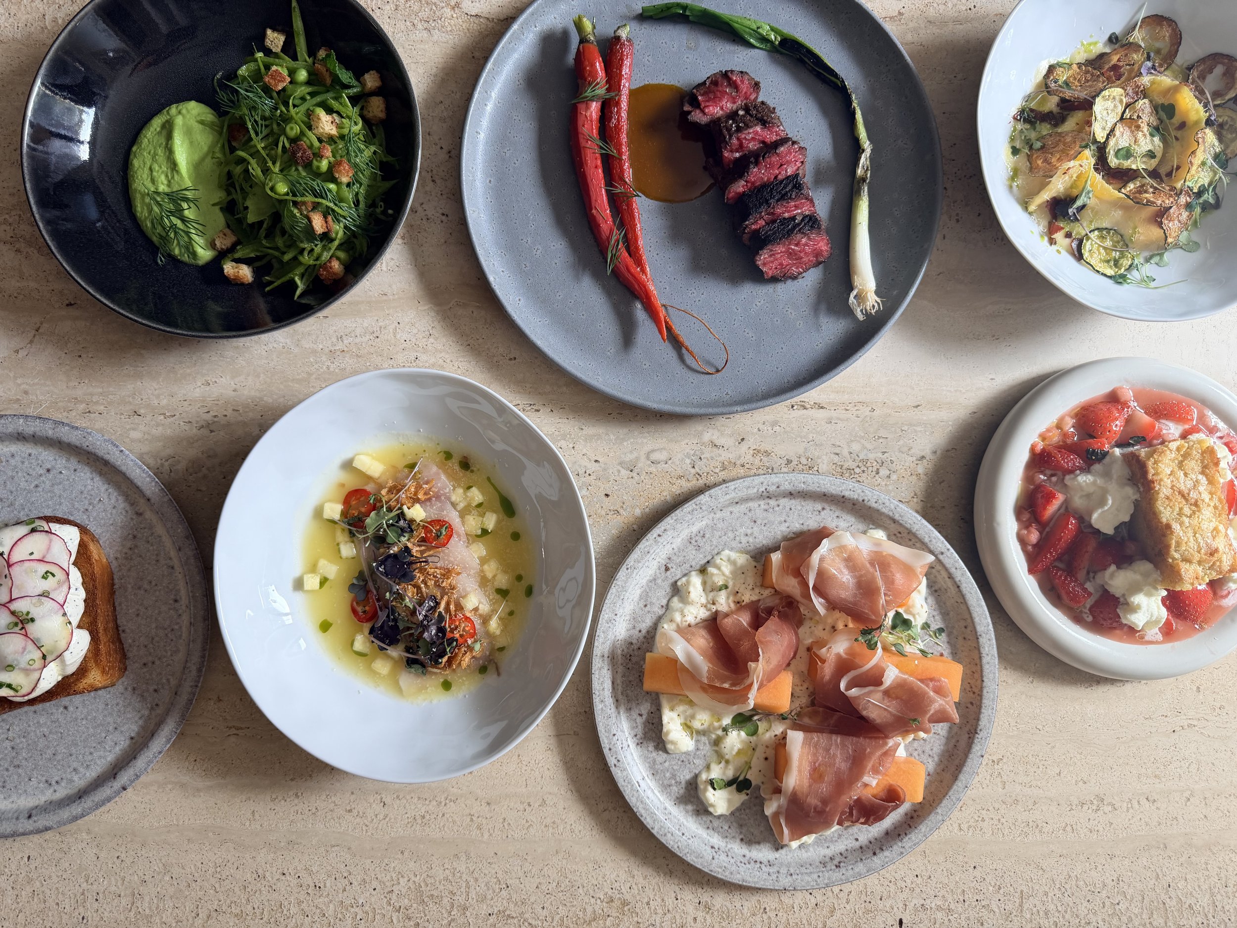 A variety of dishes on a wooden table, including a salad with peas and croutons, a plate of sliced beef with roasted red carrots and a green onion, a bowl of soup with herbs, a plate with prosciutto and cantaloupe, a slice of toast with radishes, and a dessert with strawberries.