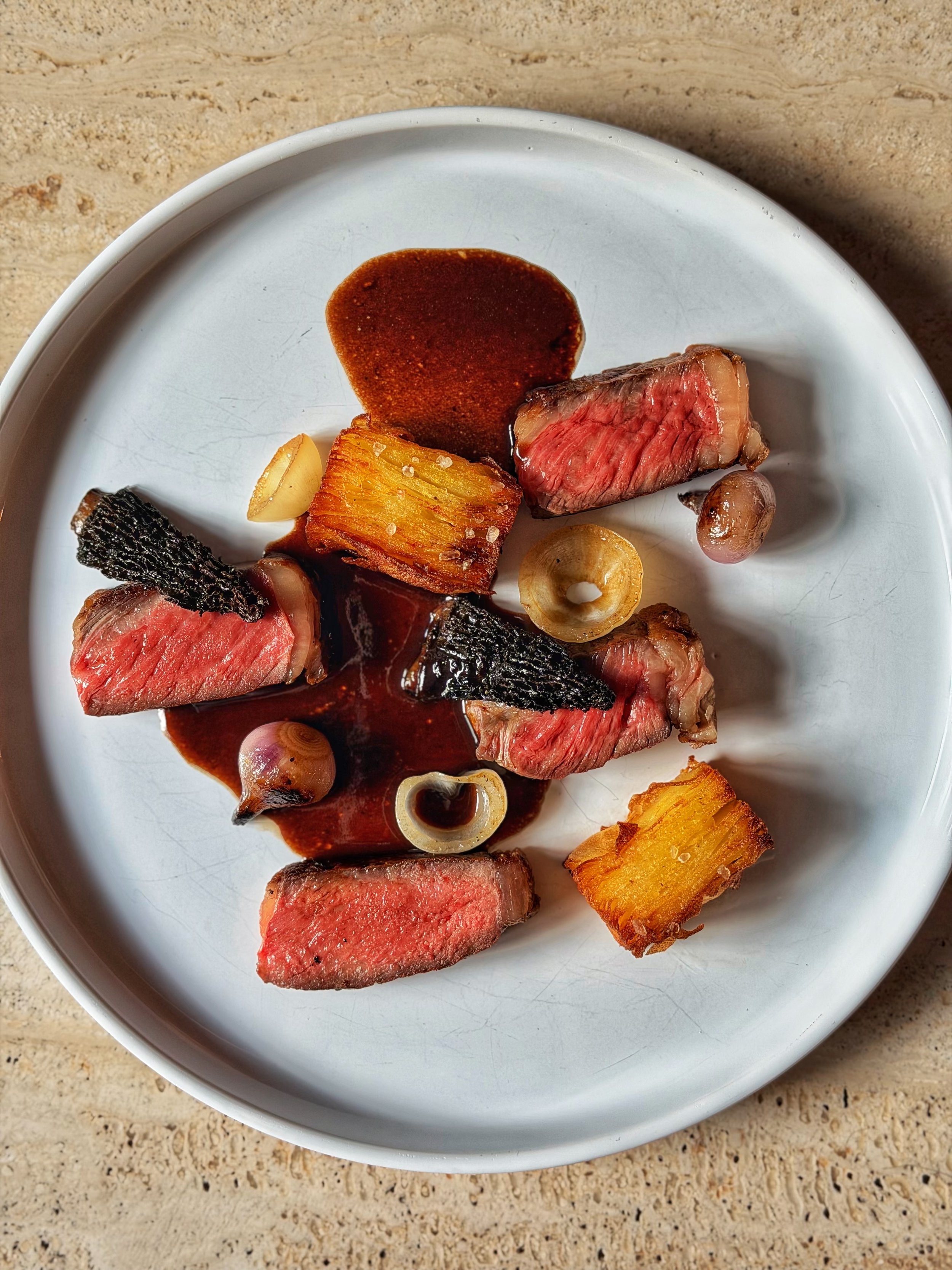 Slices of medium-rare steak with seared crust, accompanied by roasted potatoes, garlic cloves, pearl onions, and a small amount of brown sauce on a white plate.