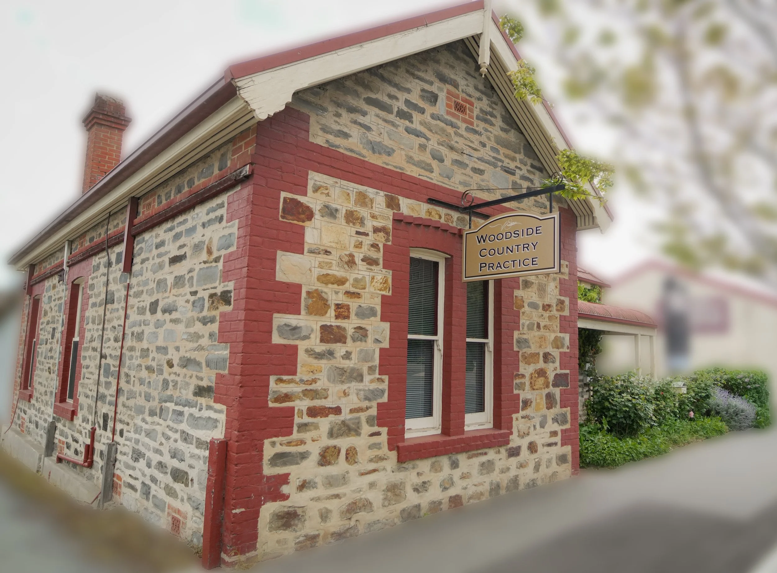 A stone building with red brick accents and a red roof, featuring a sign that reads "Woodside Country Practice," surrounded by greenery and trees.