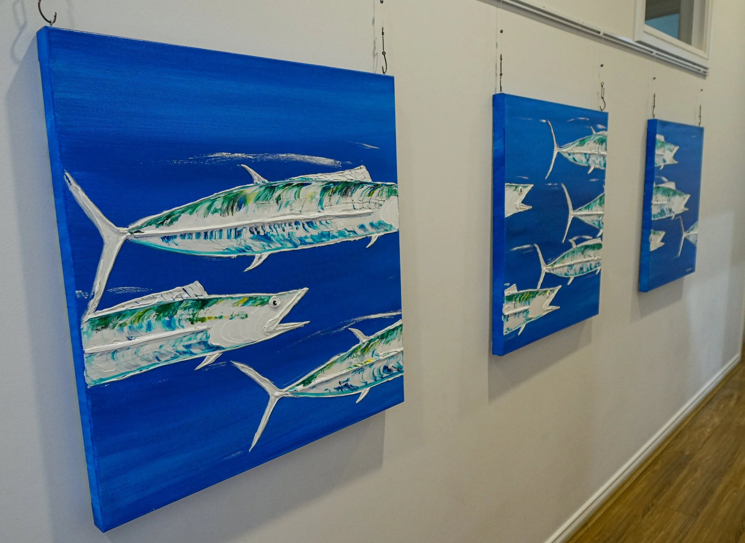 Three paintings of fish with a blue background hanging on a white wall in an art gallery.