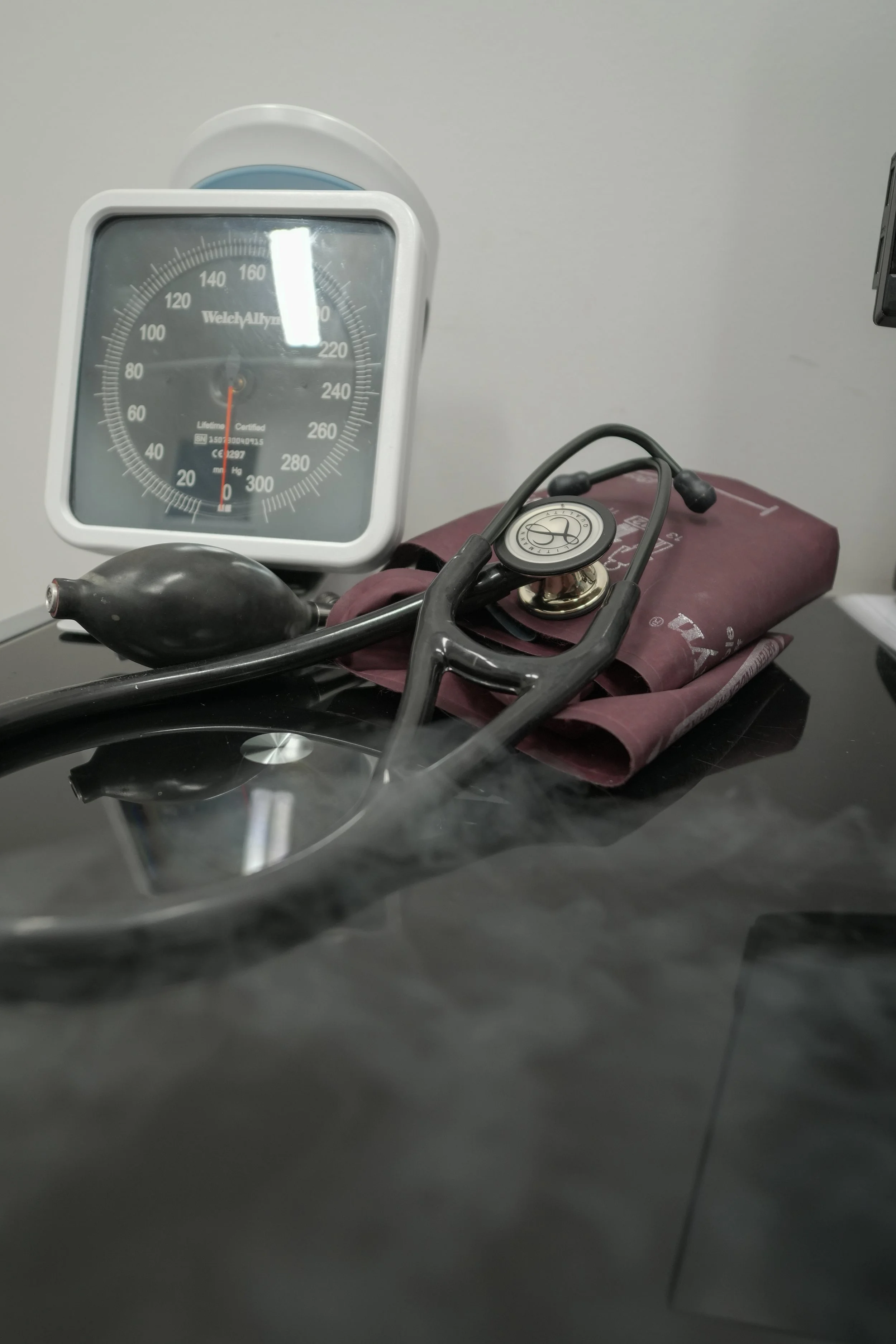 Medical equipment on a black surface including a blood pressure cuff with a rubber bulb, a stethoscope, and a wall-mounted sphygmomanometer gauge.