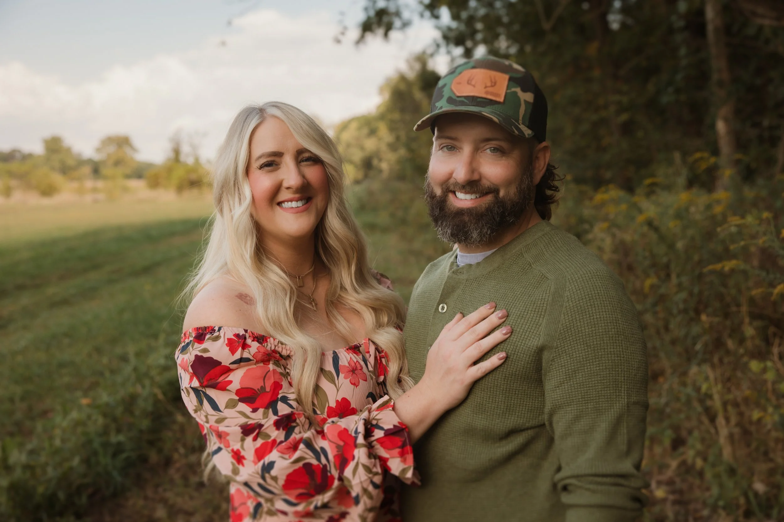 A smiling couple standing outdoors in a grassy area with trees in the background. The woman has long blonde hair, wearing a floral off-shoulder dress. The man has a beard, wearing a camouflage cap and a green sweater. The woman has her hand on the man's chest.