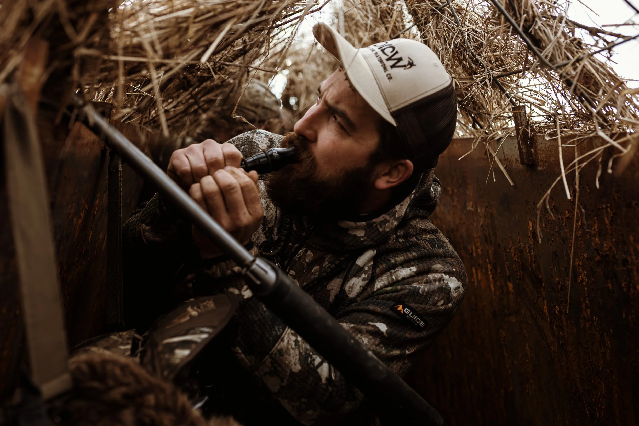 A man with a beard wearing a baseball cap and camouflage clothing is kneeling inside a trench, using a tool to dig or scrape soil and debris.