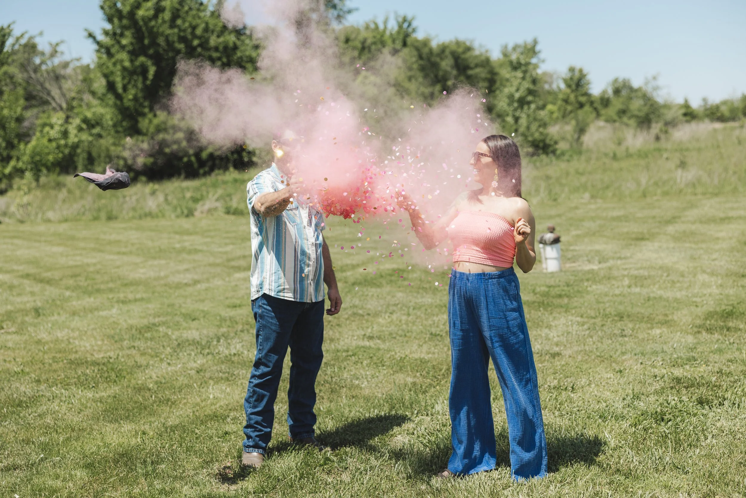 Two people playing with colored powder in an open grassy field, with trees in the background. One person is a woman wearing a pink strapless top and blue loose pants, and the other is a man wearing a striped shirt and jeans. The woman is throwing pink powder at the man, creating a cloud of pink and multicolored powder.