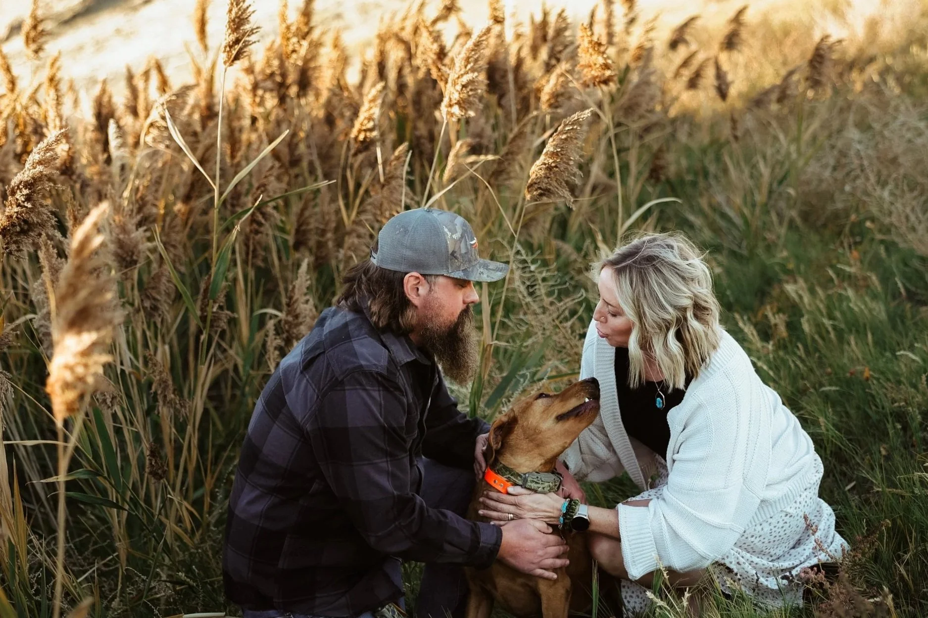 A man with long hair and a beard wearing a cap and a woman with blonde hair in a white sweater sitting in a field of tall grass and wild plants, petting a brown dog with a collar.